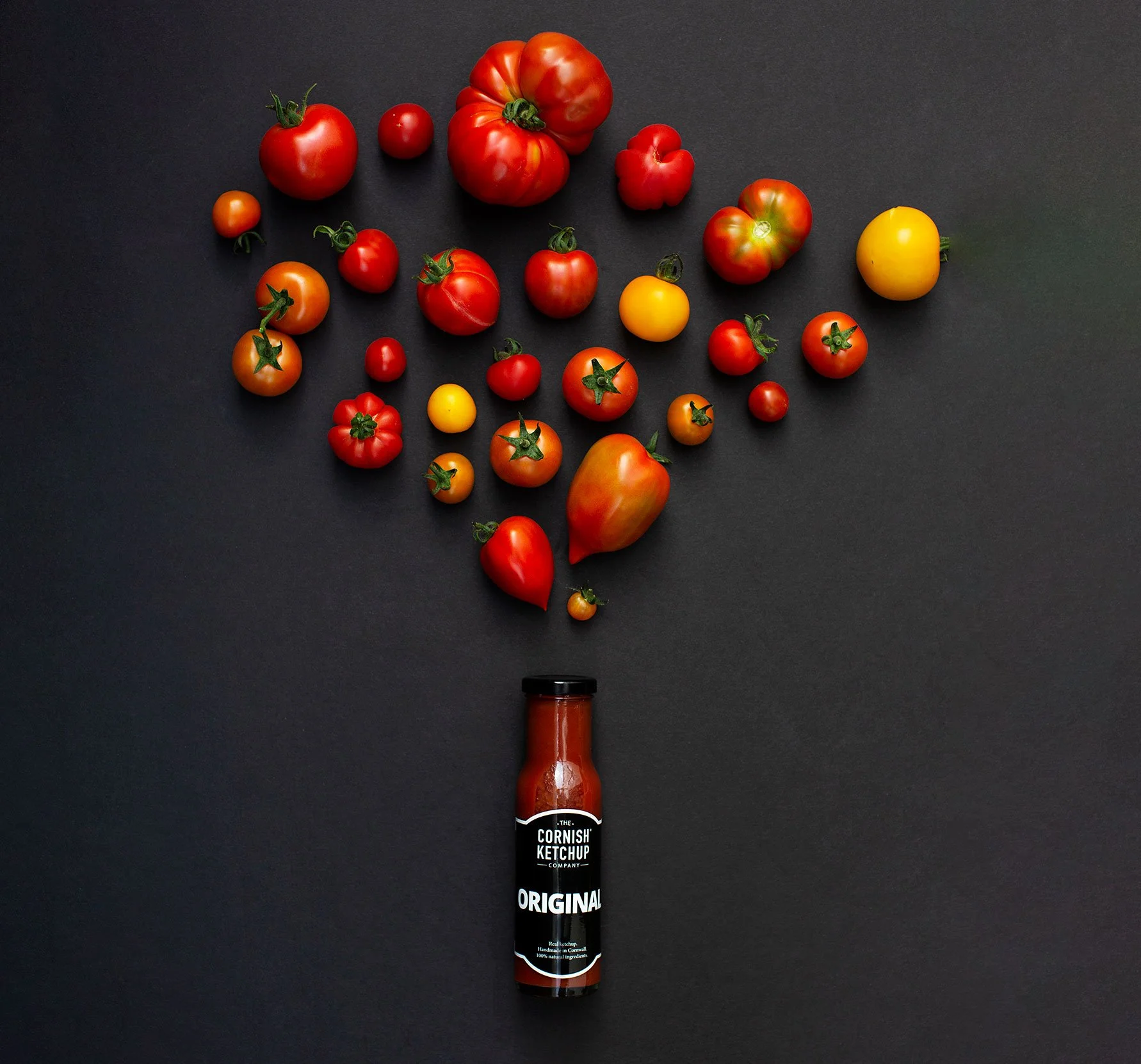 A bottle of Cornish ketchup on a black background with various heritage big and small tomatoes arranged above it.