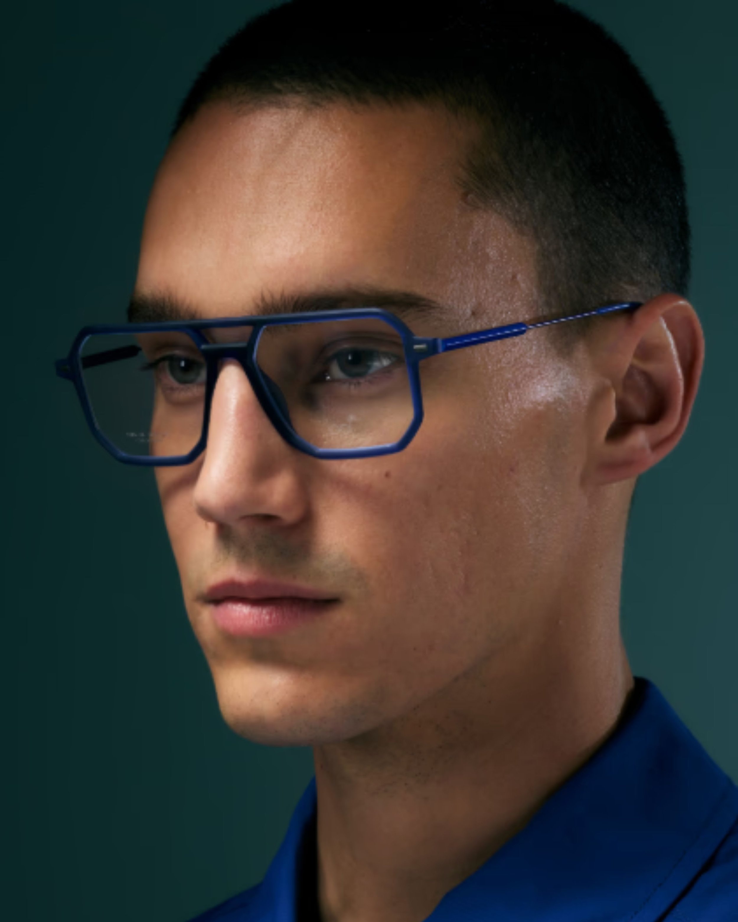 Close-up of a young man with short dark hair, wearing unique blue glasses from lool eyewear, against a dark background.