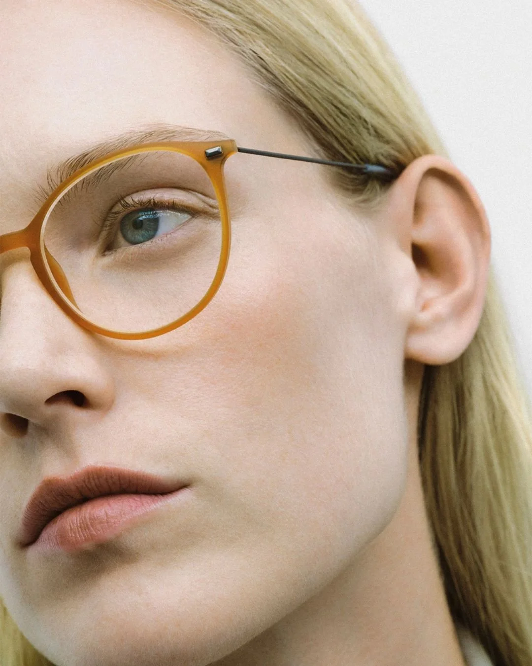 Close-up of a woman's face, with blonde hair, wearing orange-rimmed glasses, showing one eye and part of her lips.