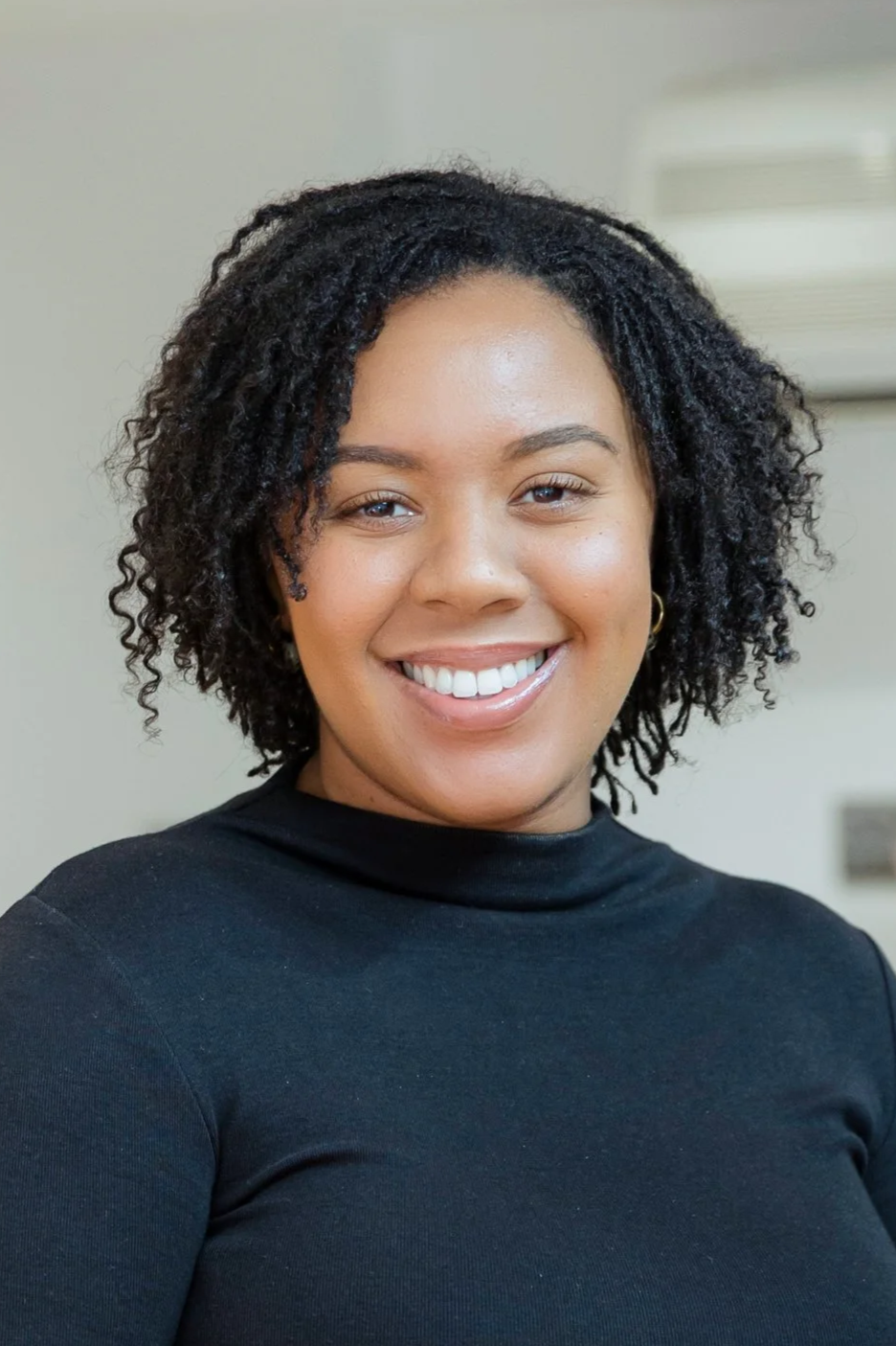 Portrait of a smiling woman with curly black hair, wearing a black top, indoors.