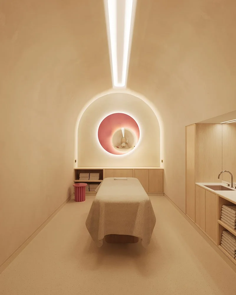Minimalist massage room with an off-white color scheme, a massage table covered with a beige sheet, a pink decorative stool, a shelf with folded towels, and abstract wall art illuminated by soft lighting.