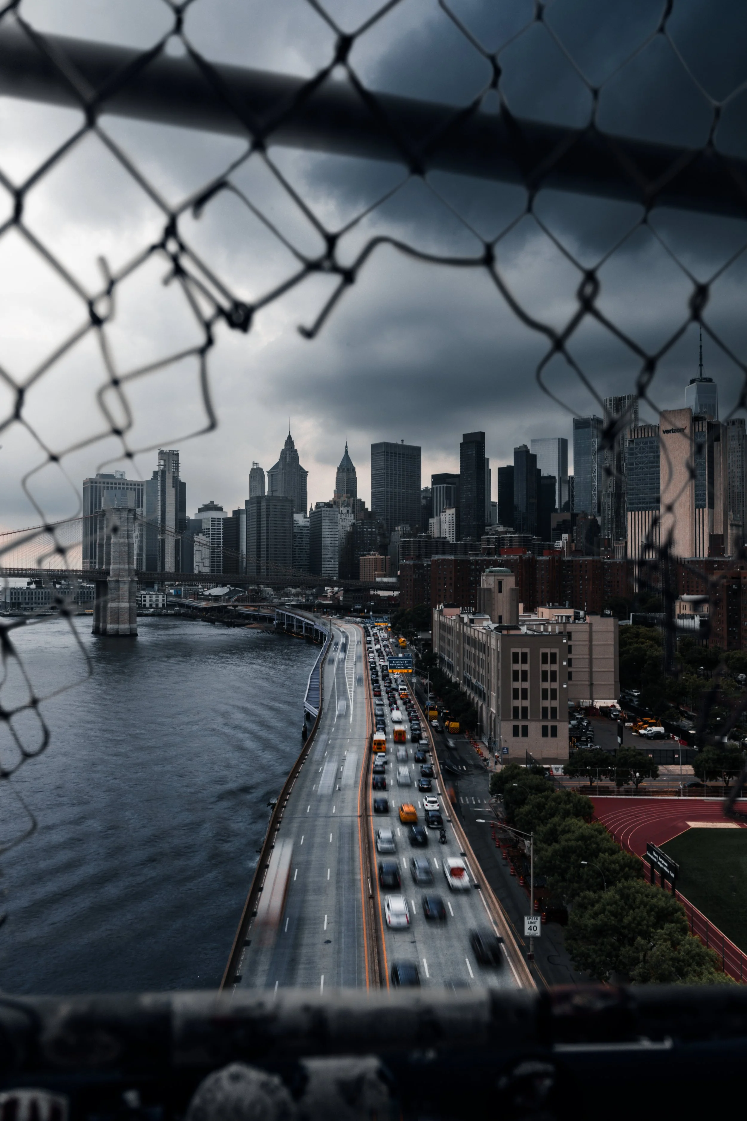View of a city's skyline with tall skyscrapers, a bridge over the water, and a busy road with traffic, seen through a chain-link fence on a cloudy day.