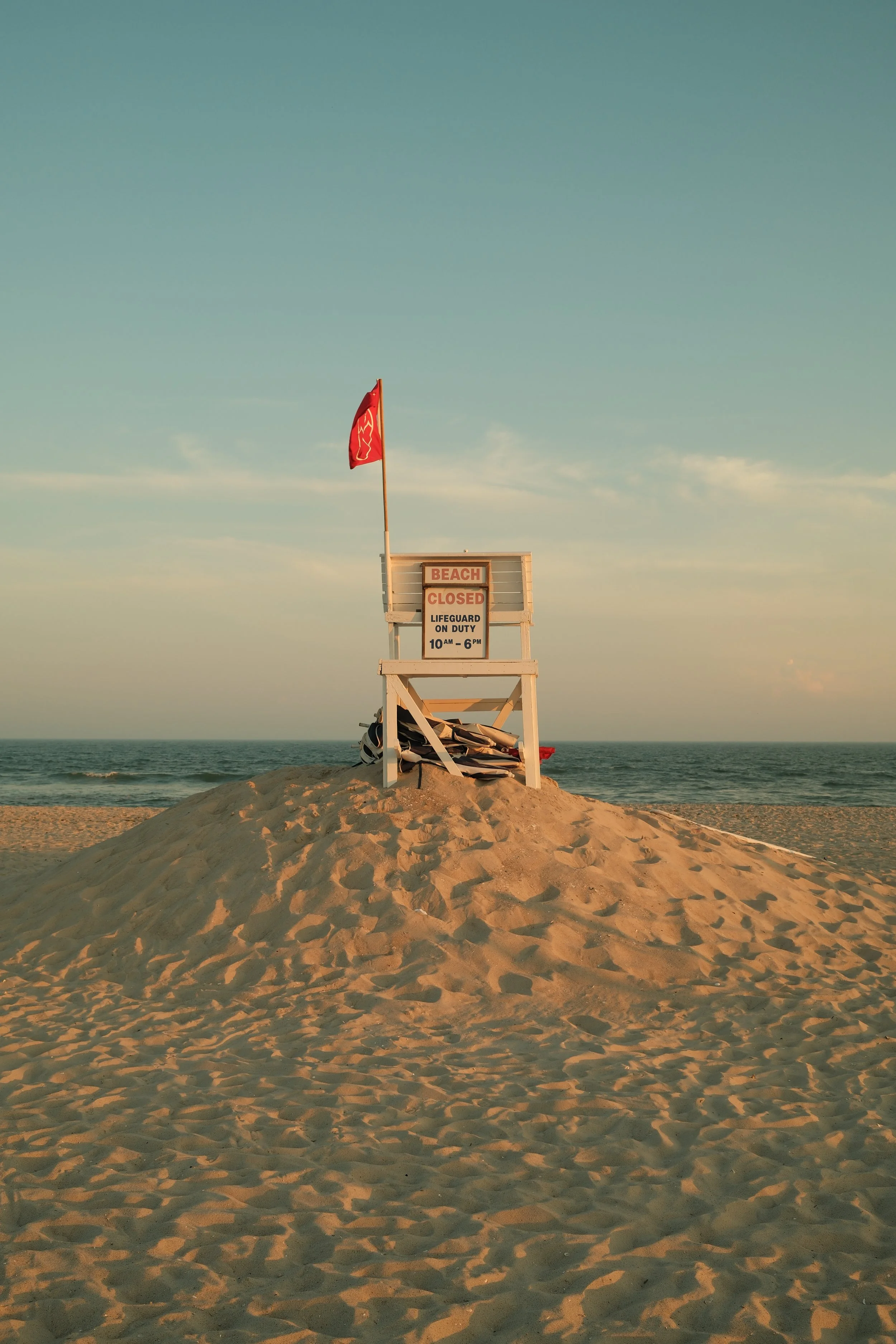 A lifeguard tower on a sandy beach with a red flag, sign indicating beach is closed and lifeguard on duty from 10 am to 6 pm, overlooking the ocean at sunset.