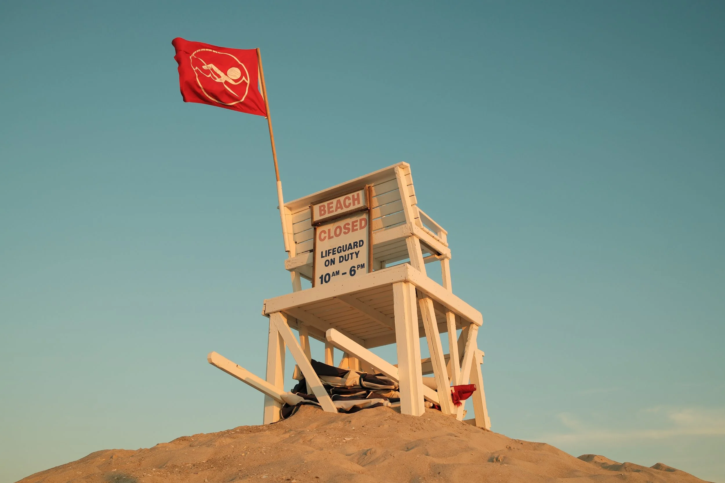 Empty white lifeguard tower on a sandy beach with a red flag and a sign that reads "Beach Closed, Lifeguard on Duty 10 am - 6 pm," under a clear sky.
