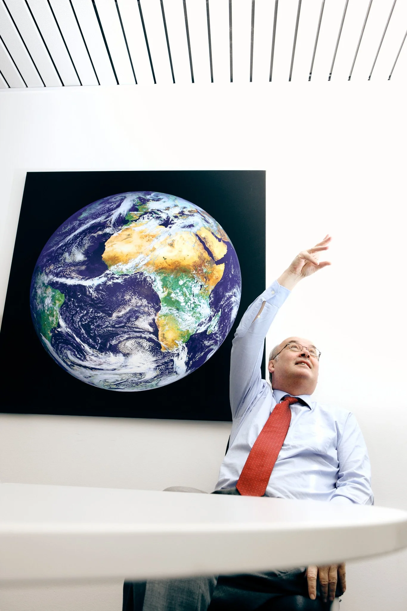 Ein Mann sitzt in einem Büro vor einem Bild der Erde, hebt den Arm zur Präsentation.