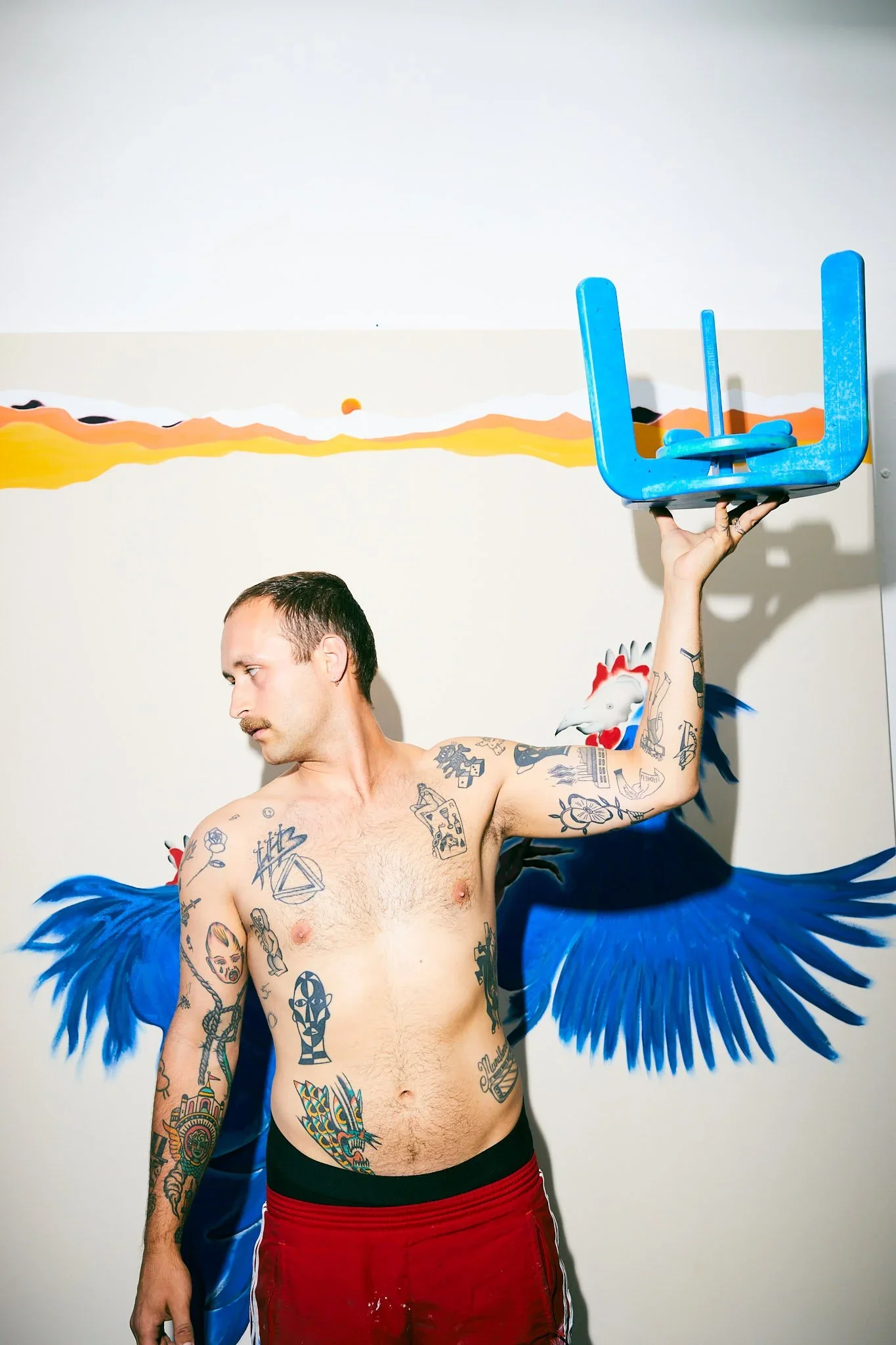 Ein nackter Mann mit Tattoos hält einen blauen Hocker in der Hand, vor einer Wand mit Wandmalerei eines blauen Vogels mit ausgebreiteten Flügeln.