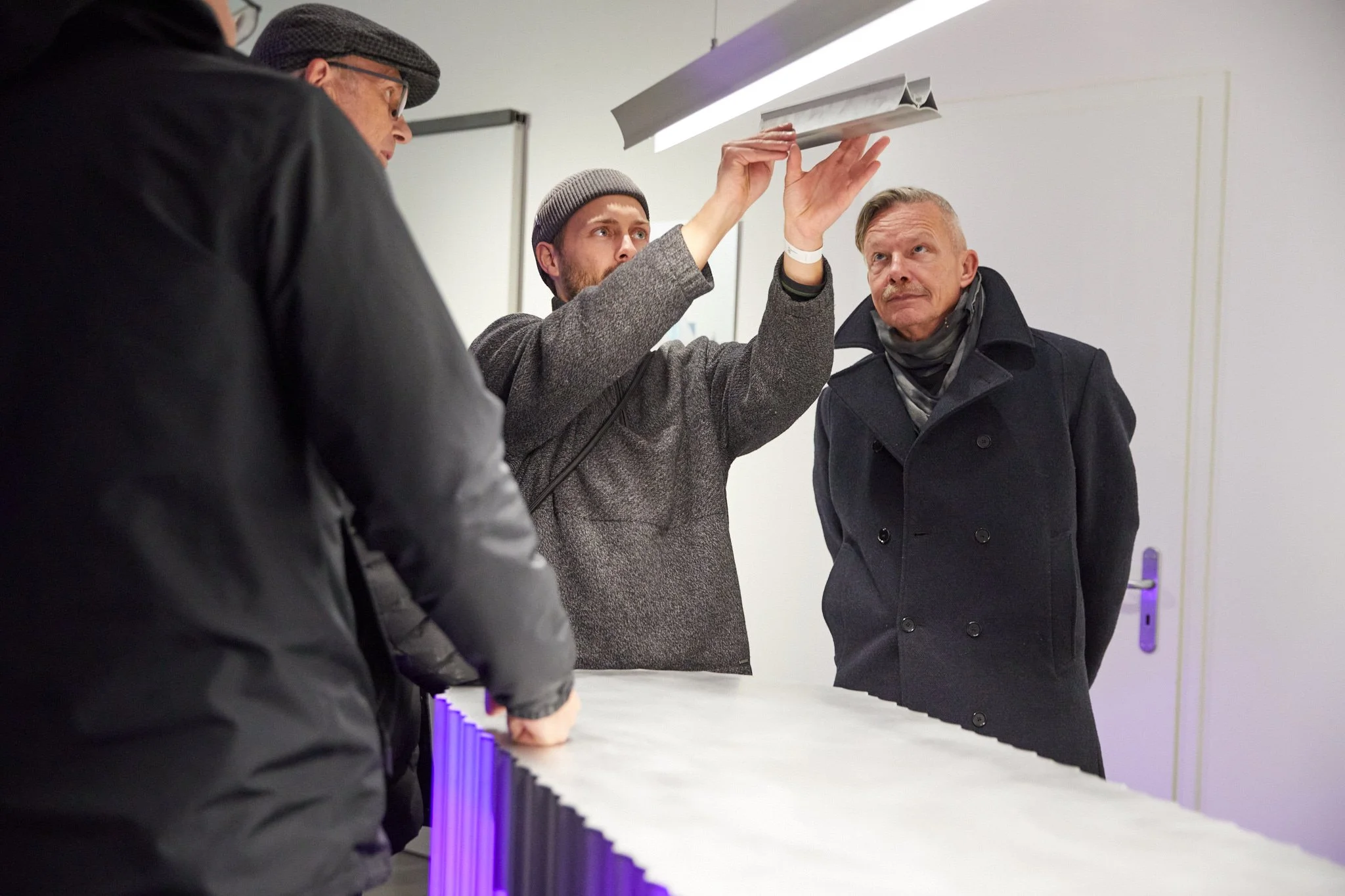 Drei Männer stehen um einen Tisch, einer erklärt etwas, während die anderen beiden zuhören. Der Raum ist modern und gut beleuchtet.