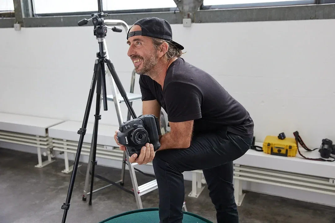 Ein lächelnder Mann mit dunklem T-Shirt, schwarzer Kappe und Kamera in der Hand, fotografiert in einem Raum mit großen Fenstern und Equipment auf der Bank.