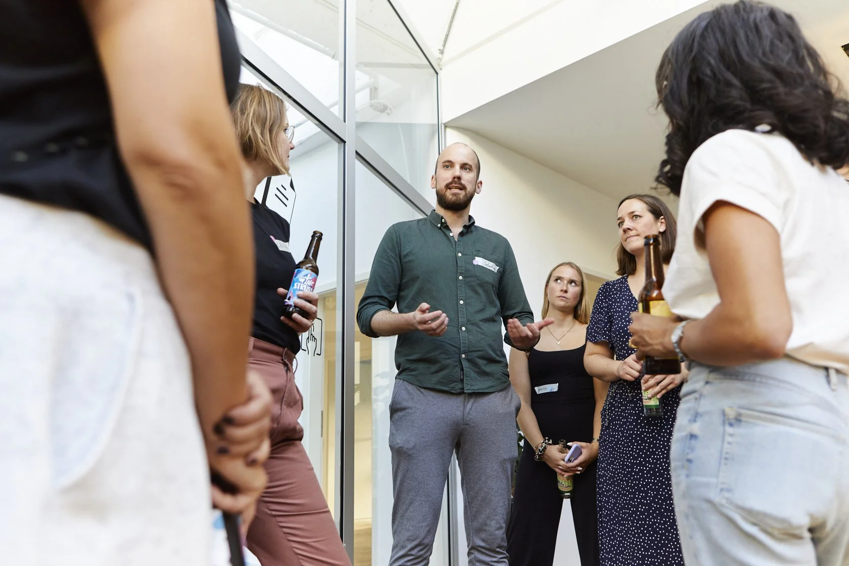 Eine Gruppe von fünf Frauen und einem Mann im Gespräch in einem modernen Innenraum, die Frauen halten Getränke. Der Mann wirkt, als würde er etwas erklären.