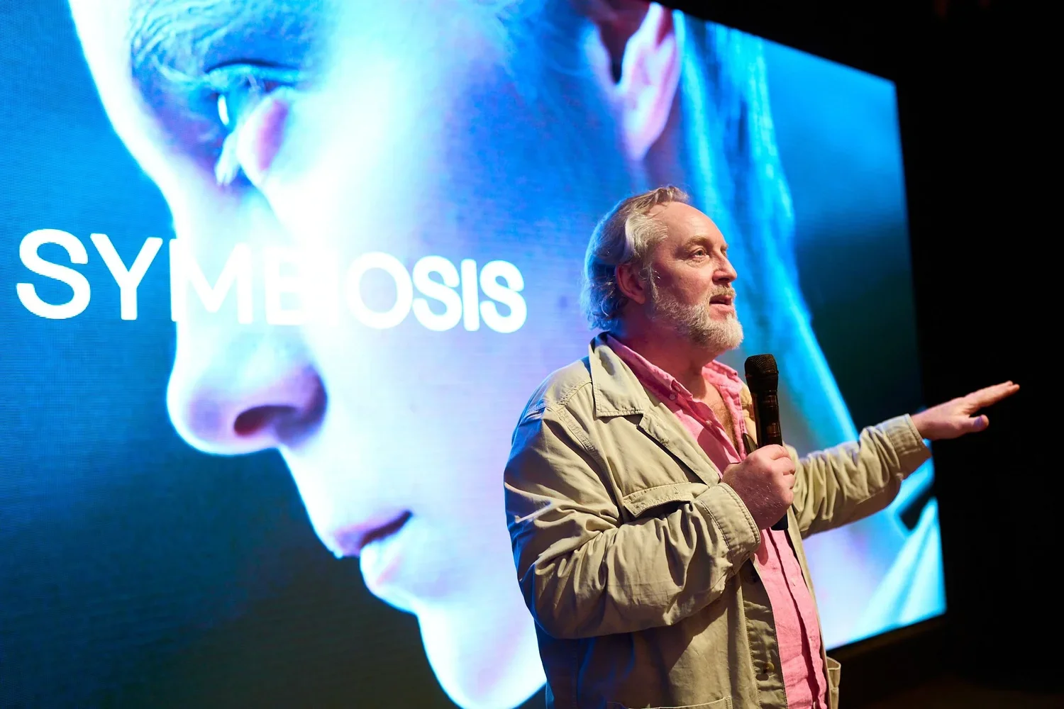 Ein Mann mit grauem Haar und Bart hält ein Mikrofon in der Hand und zeigt auf eine große Leinwand im Hintergrund, auf der das Wort 'SYMBOLISATION' neben einem facialen Porträt einer Person in Blau- und Lilatönen zu sehen ist.
