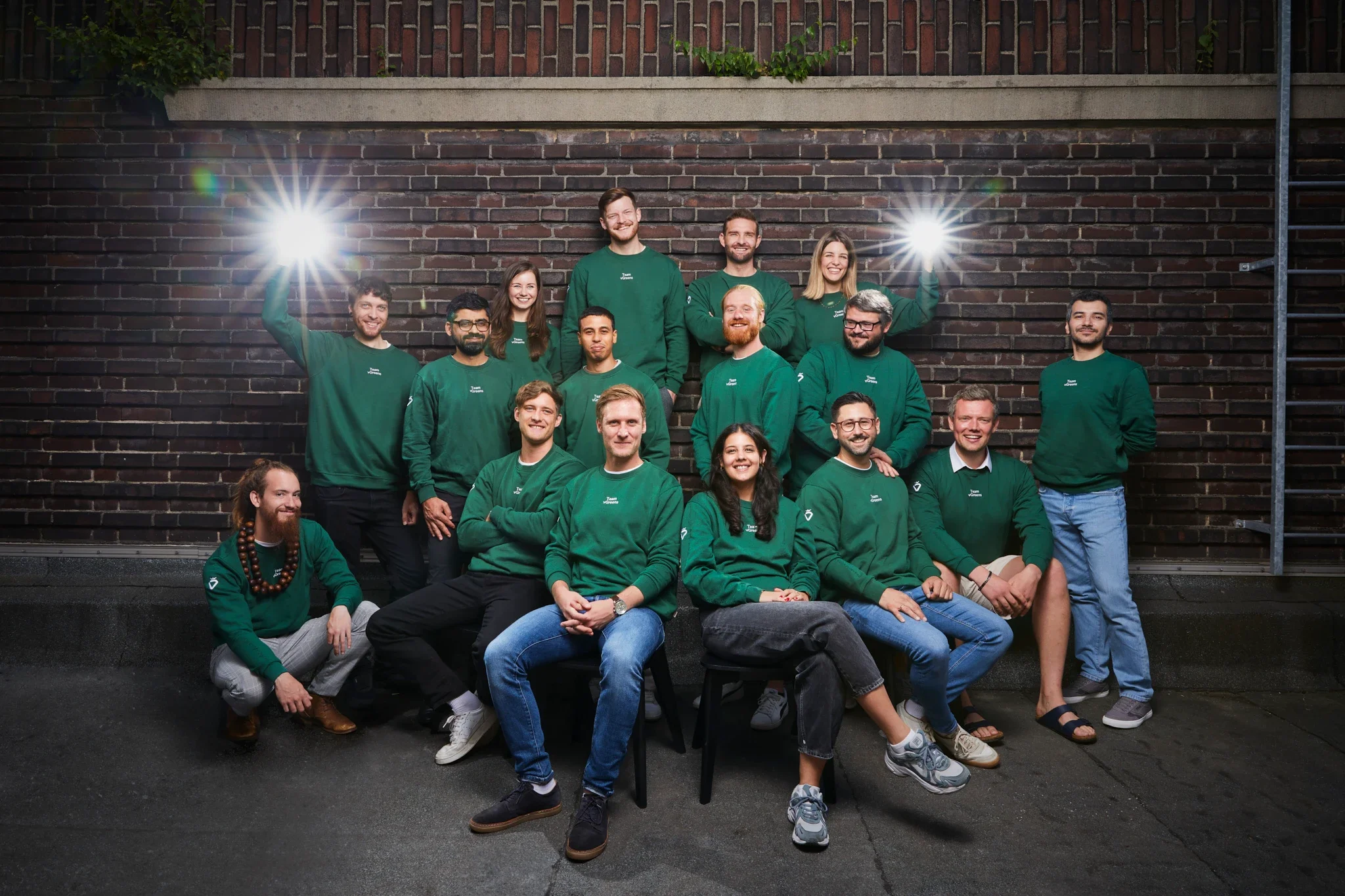 Eine Gruppe junger Menschen trägt grüne Sweatshirts und posiert für ein Gruppenfoto vor einer dunklen Backsteinmauer bei Nacht, zwei von ihnen halten Blitzlichter. Sie sind freundlich und lächeln.