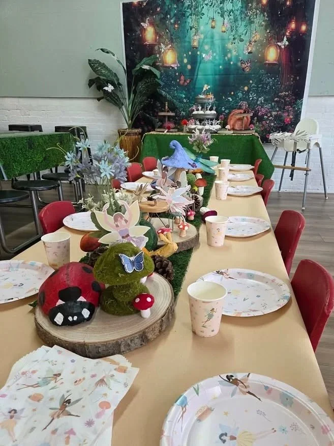 Decorated party room with a Buzz Lightyear birthday banner, themed balloons, and a long table set with plates and cups for children, including a centerpiece with greenery.