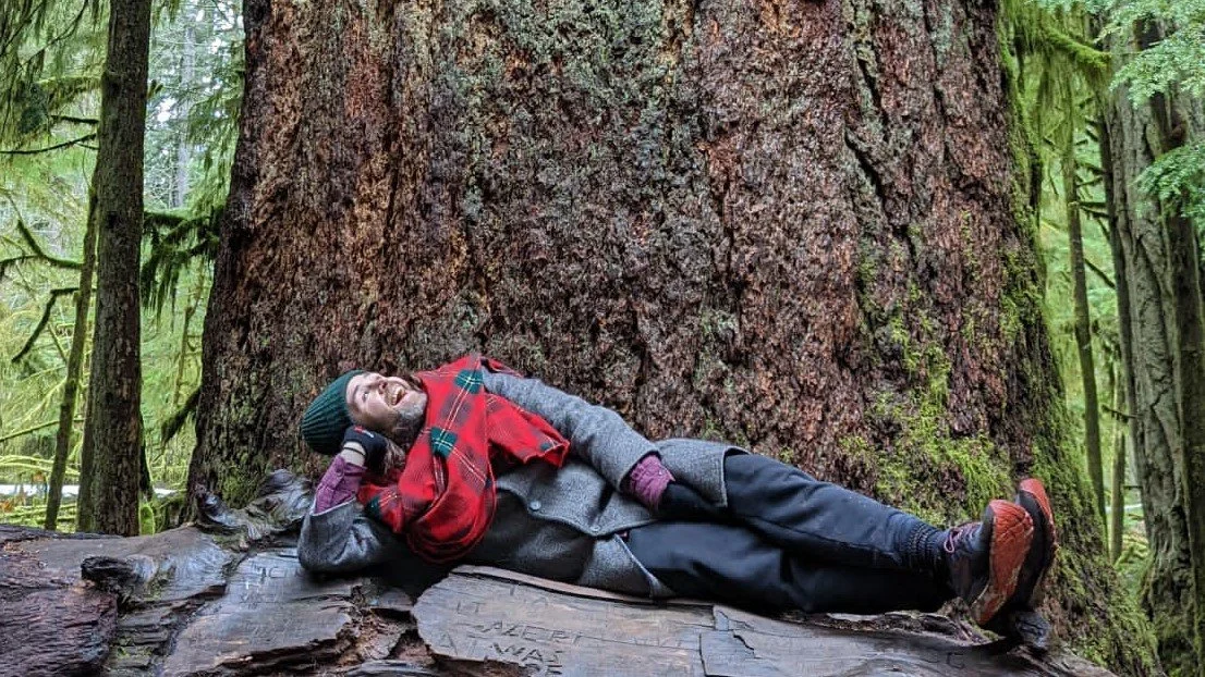 A man lying on a fallen log in front of a large tree trunk in a forest, laughing and resting.