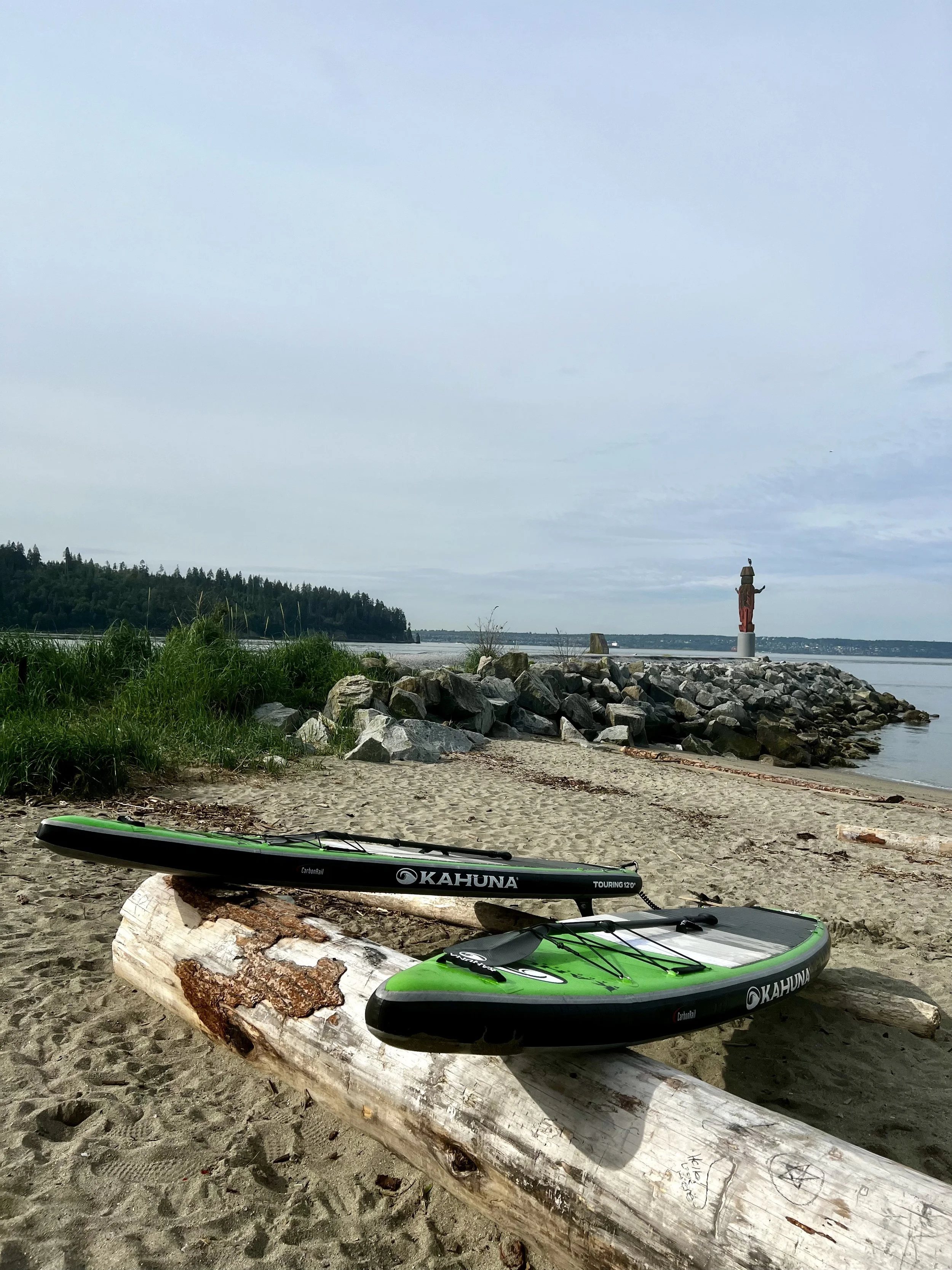 Two green and black Kahuna paddleboards resting on a log on a sandy beach with rocks near the water, a statue on a small island in the distance under a cloudy sky.