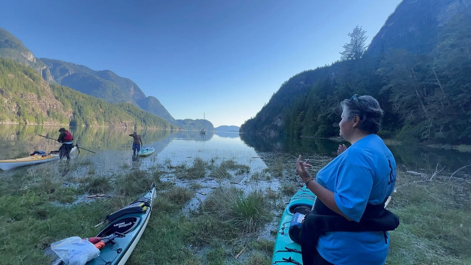 People preparing kayaks by a calm lake surrounded by mountains with dense greenery, under a clear blue sky.