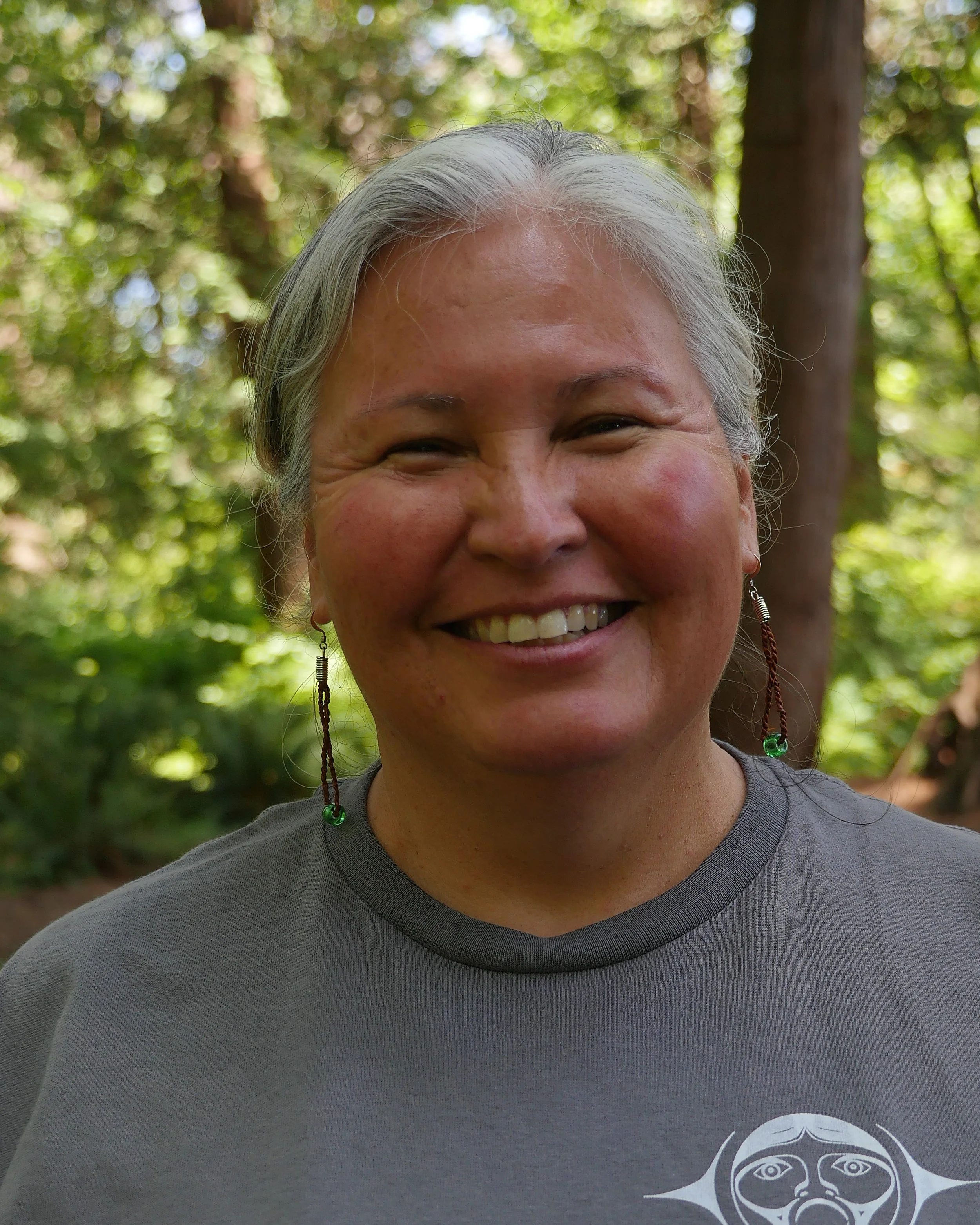 A smiling woman with gray hair outdoors, wearing earrings and a gray T-shirt with a logo that says 'Talaysay Tours' and features a stylized face.