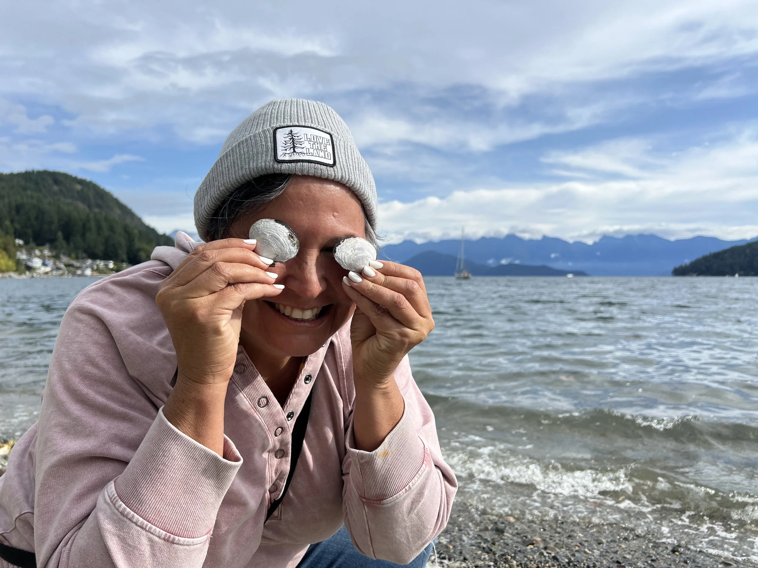 Smiling woman at the water's edge holding silver-colored rocks near her eyes, wearing a gray beanie, pink hoodie, with a mountain and lake landscape in the background.