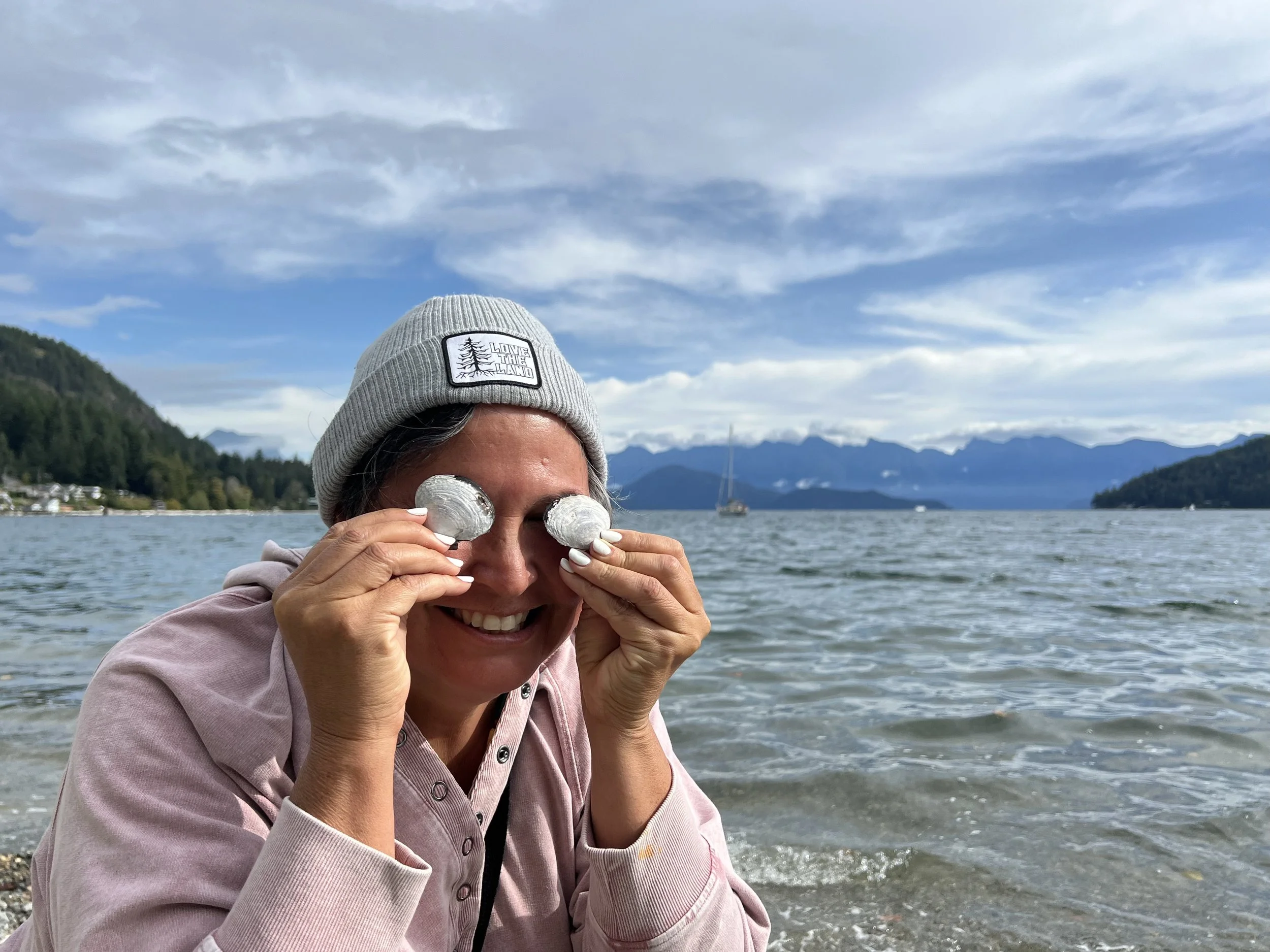 A woman smiling and holding two foil-wrapped objects over her eyes, sitting by a lake with mountains and a cloudy sky in the background.