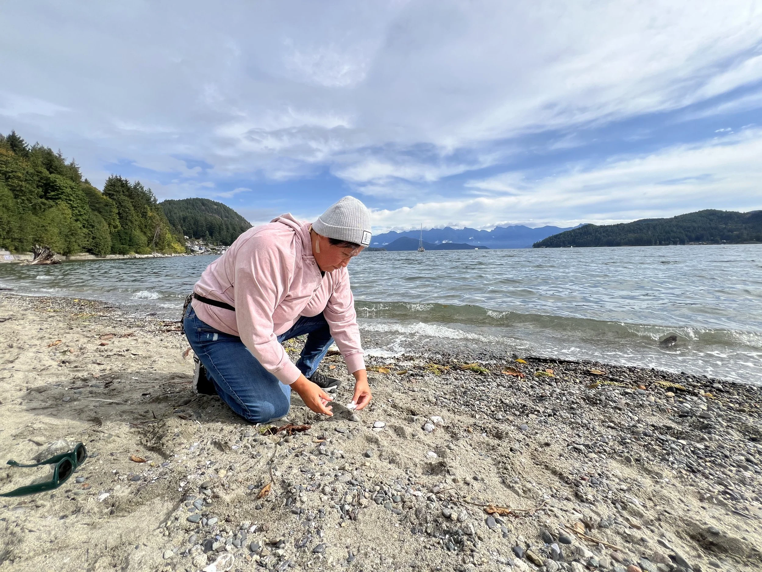 Person sitting on a sandy beach near the water, wearing a pink hoodie, gray beanie, and jeans, examining seashells with mountains and a cloudy sky in the background.