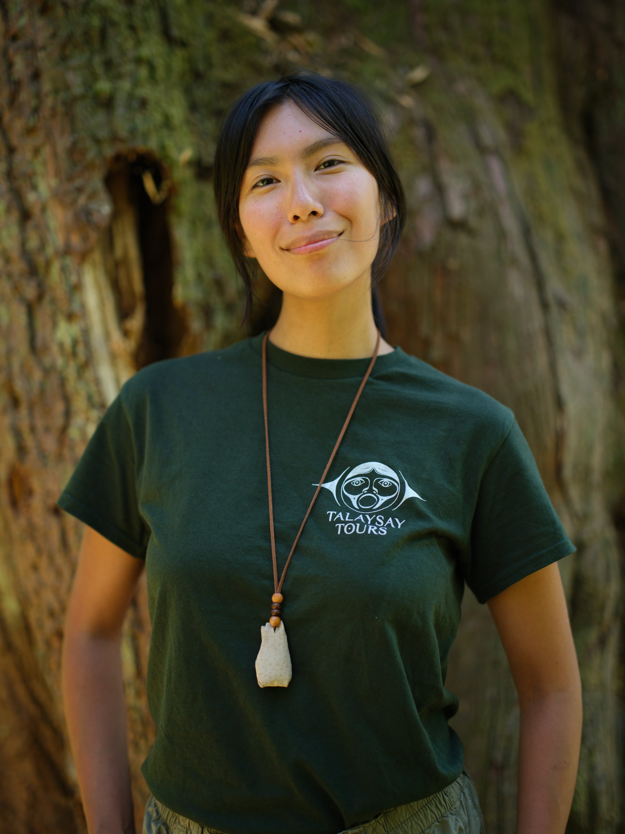 A woman wearing traditional indigenous attire, including a beaded necklace, a large pendant, and fringed clothing, sitting outdoors beside a body of water with trees in the background, smiling.