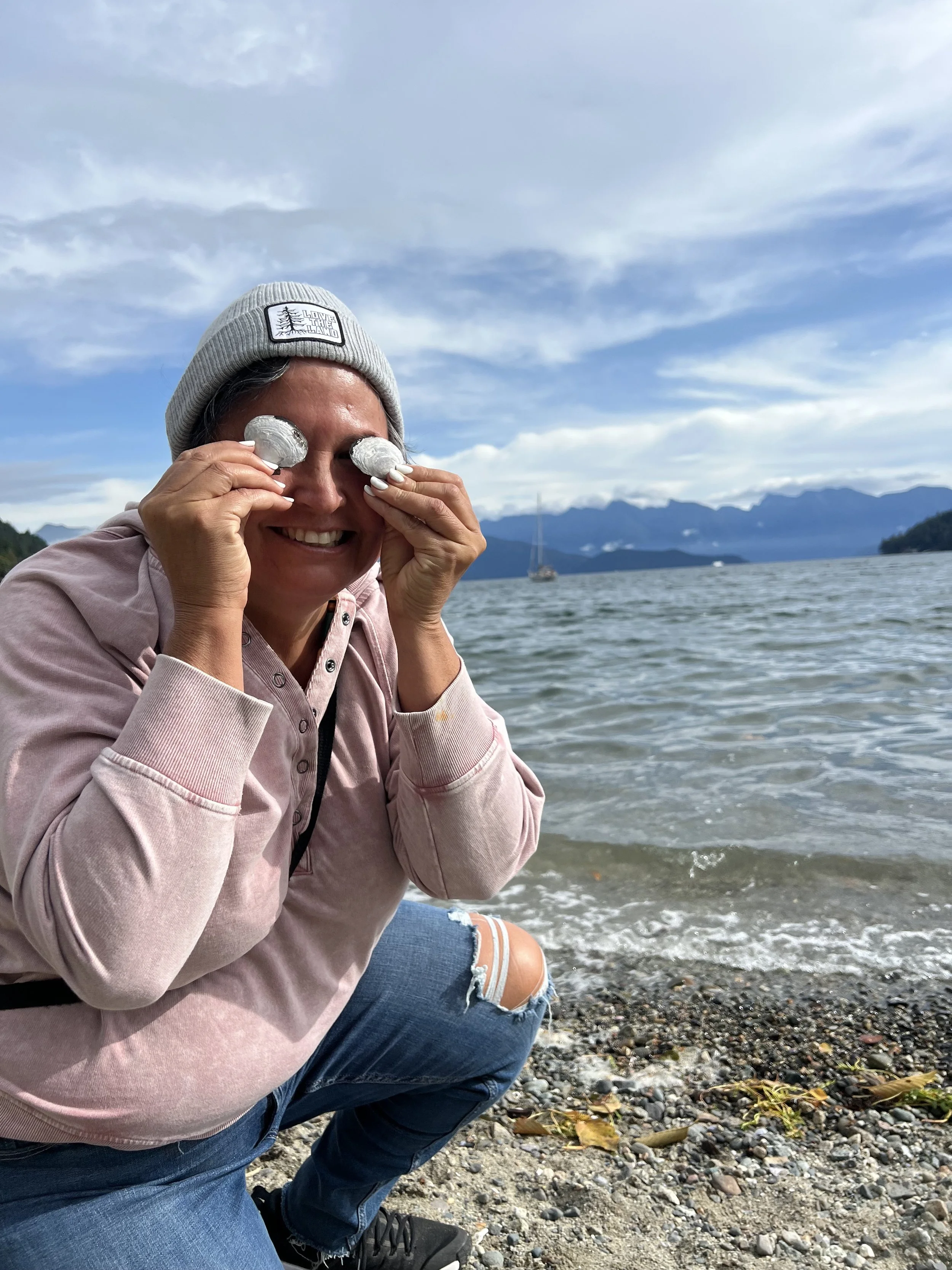 A woman with a gray beanie smiling and holding two small objects over her eyes, kneeling by a lakeshore with mountains in the background and a sailboat on the water.