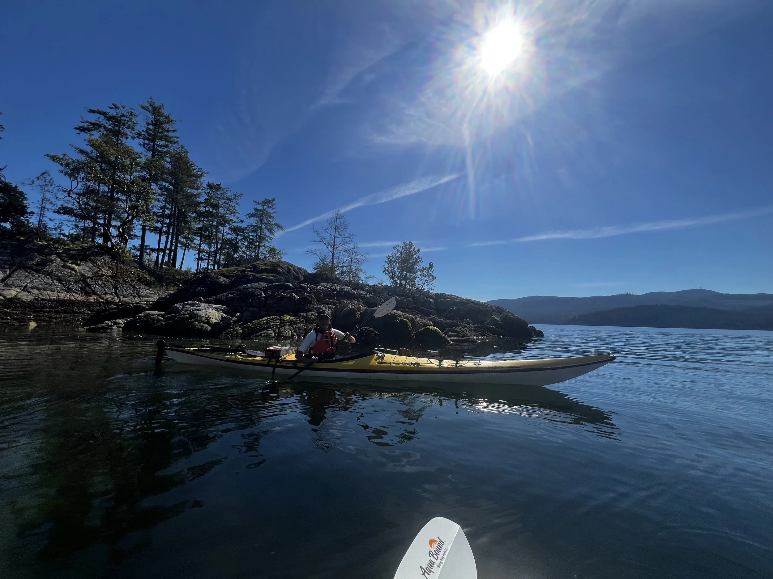 Person kayaking on a calm lake with rocky shoreline and trees under a bright sunny sky
