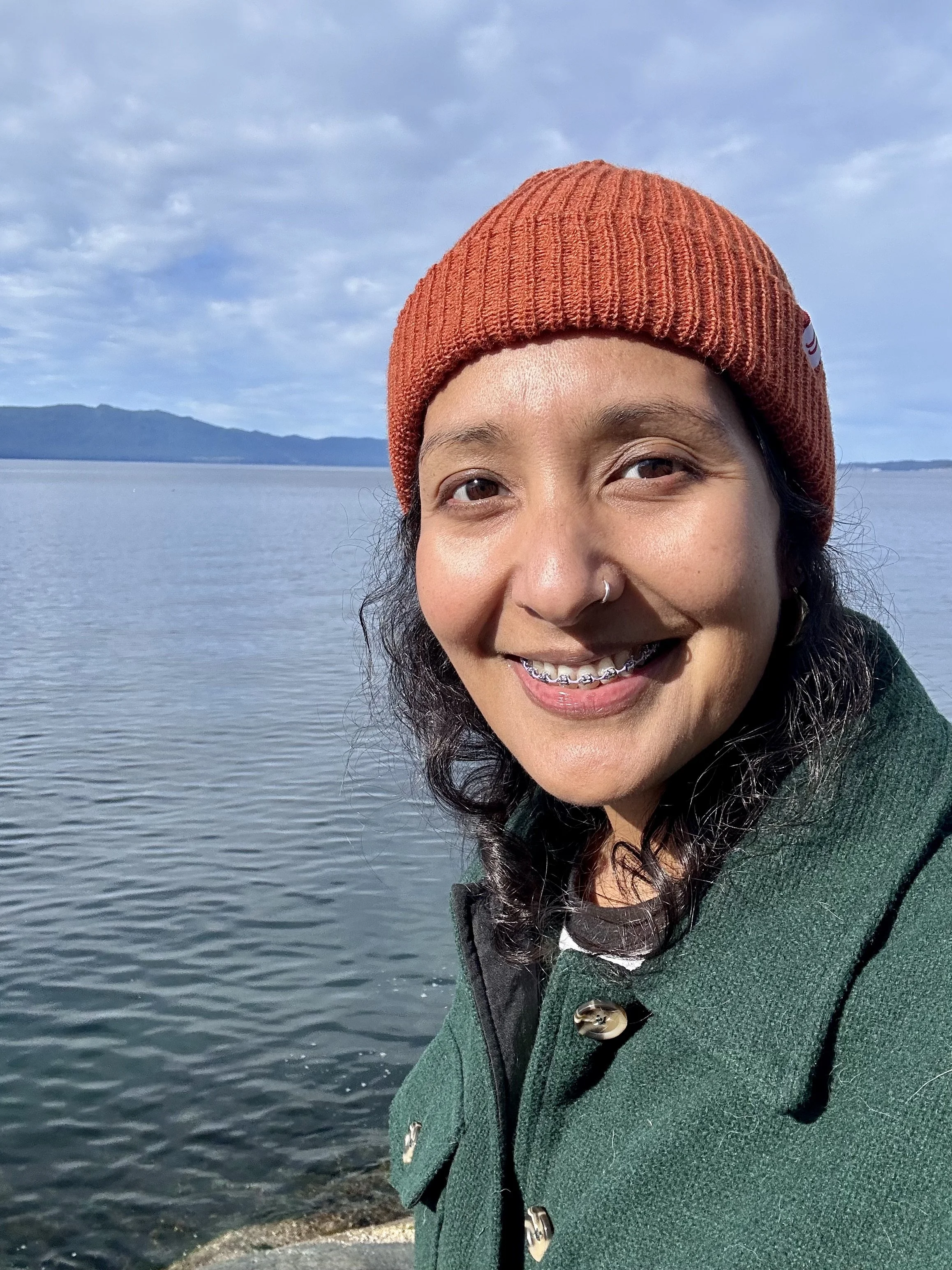 Smiling woman with long black hair, wearing an orange beanie, a nose ring, and a green coat, standing near a body of water with mountains in the background.
