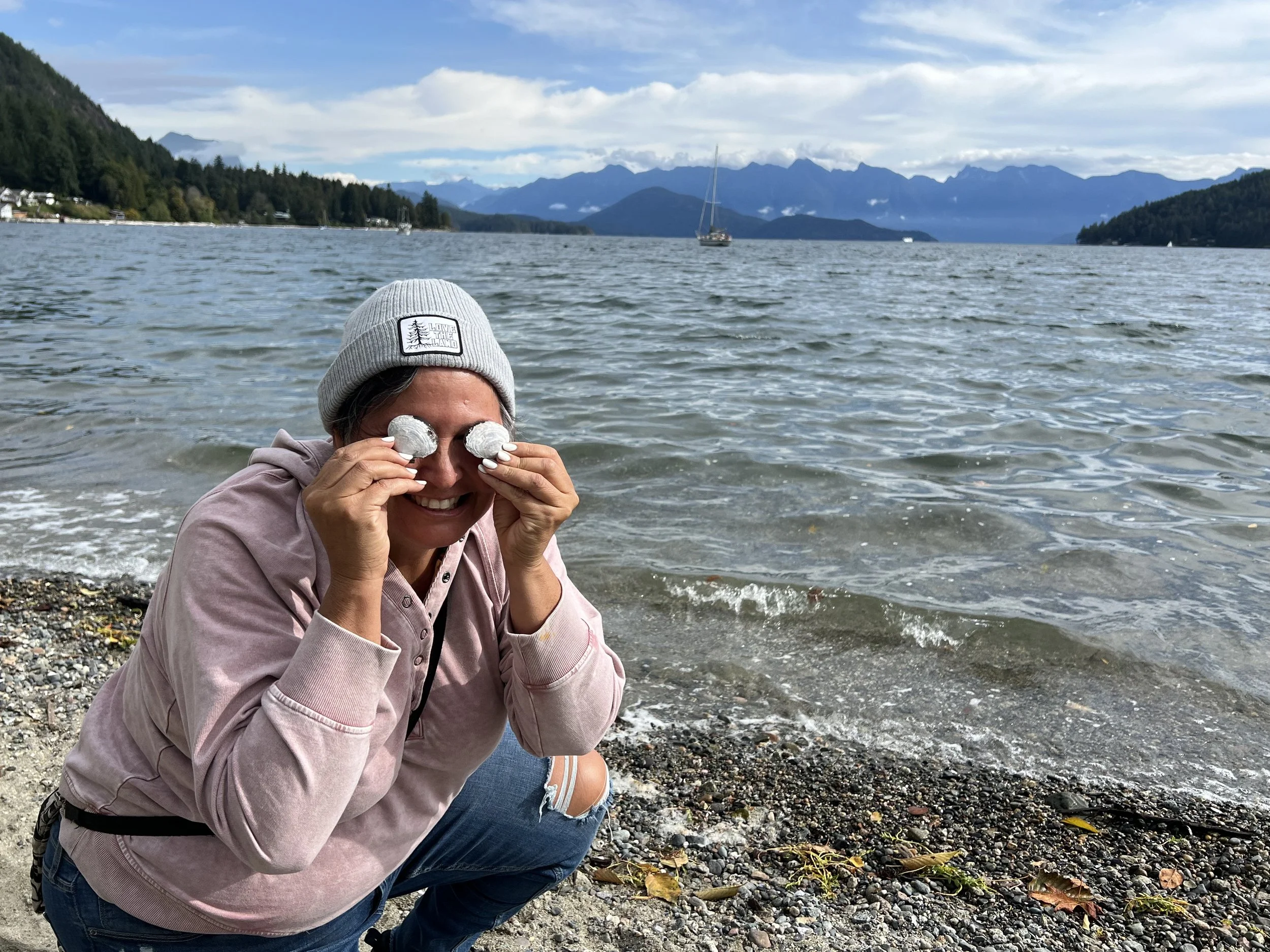 Woman squatting on a rocky beach near a lake, holding two foamy rocks in front of her eyes, smiling, with a mountain range and sailboat in the background.