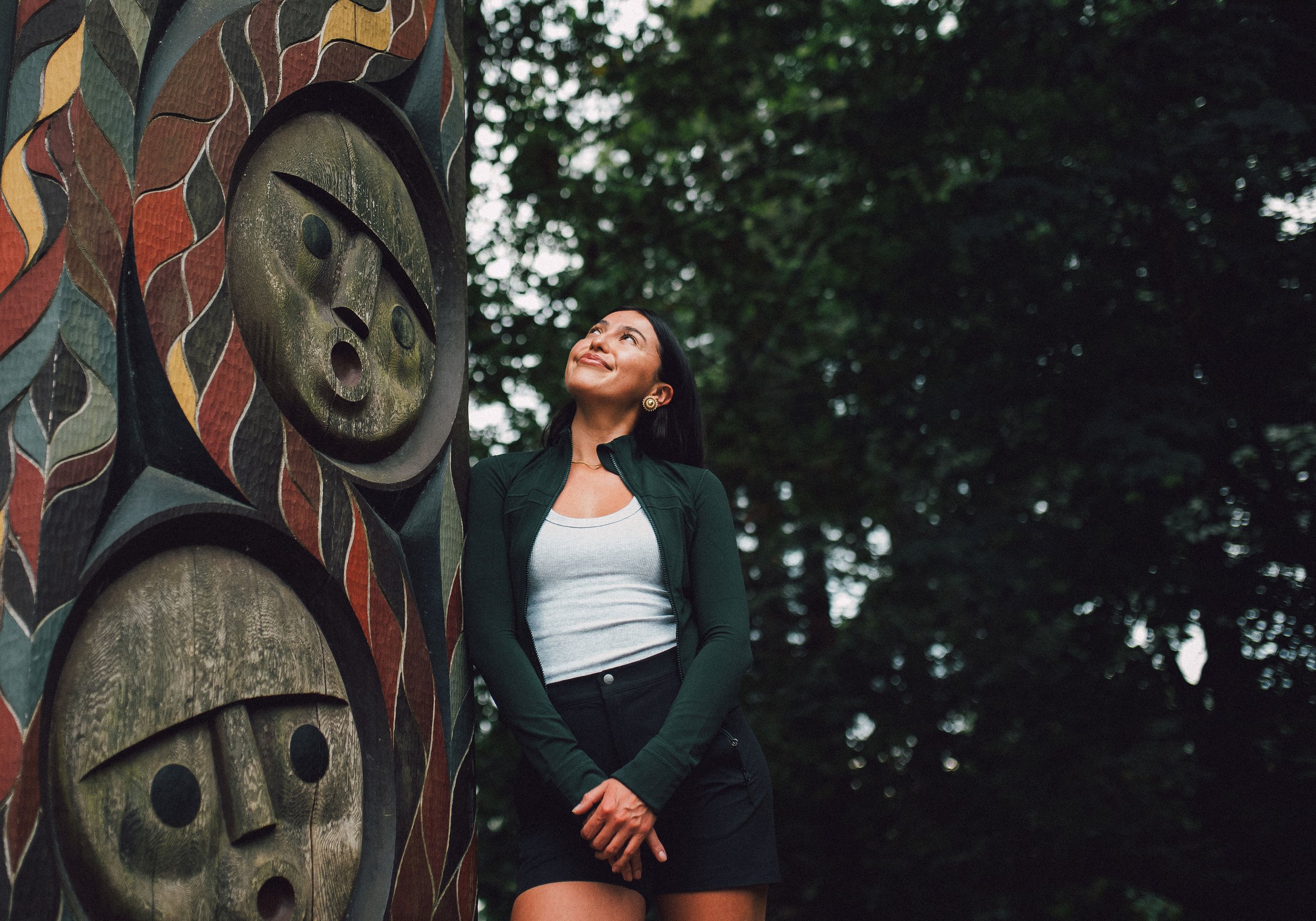 A woman with dark hair and earrings gazes upward while standing outdoors next to an art piece on a tree, surrounded by green trees and foliage.
