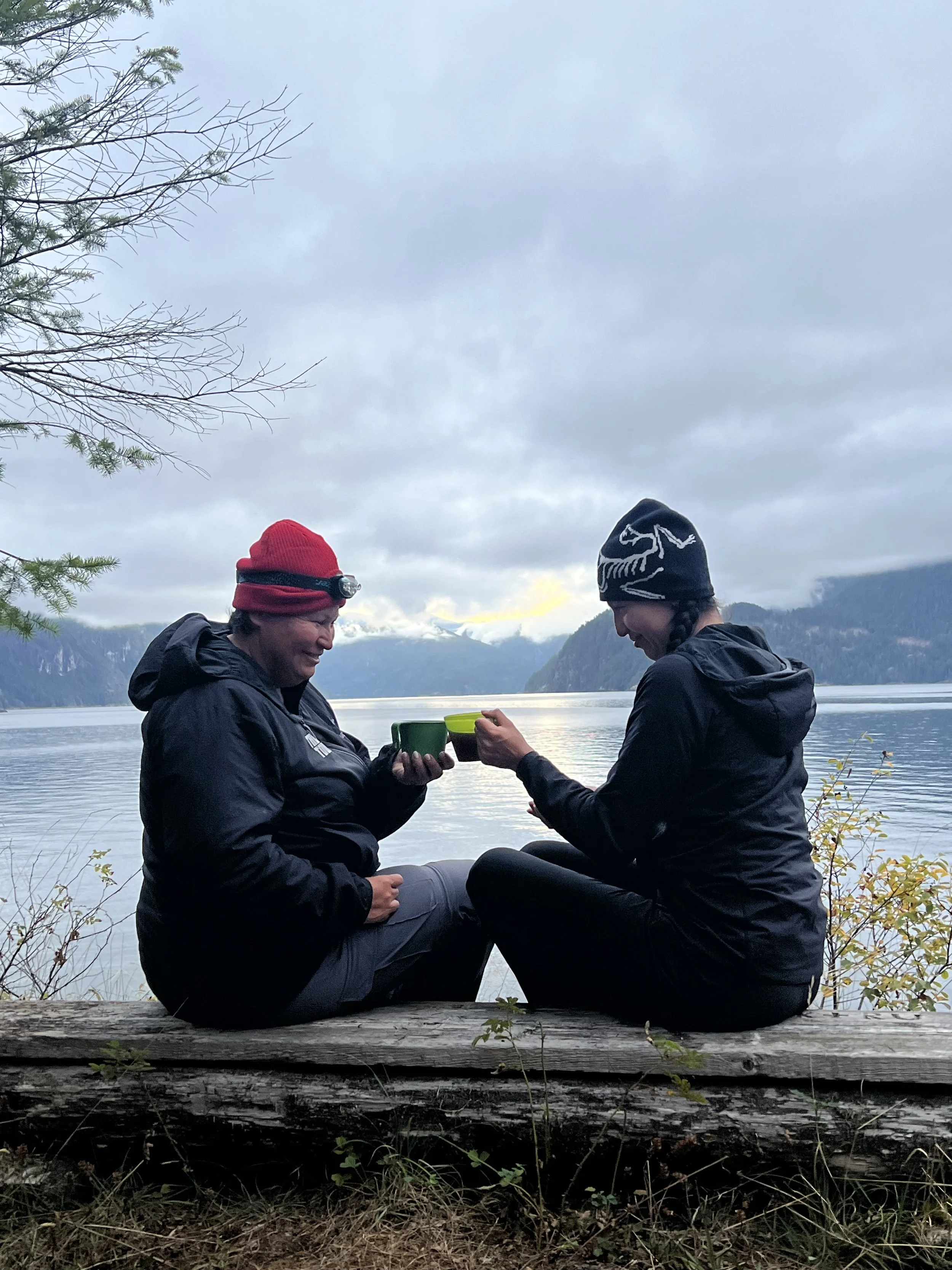 Two people sit by a lake on a large fallen log, having a drink together. They are dressed warmly in jackets and beanies, with mountains and a cloudy sky in the background.