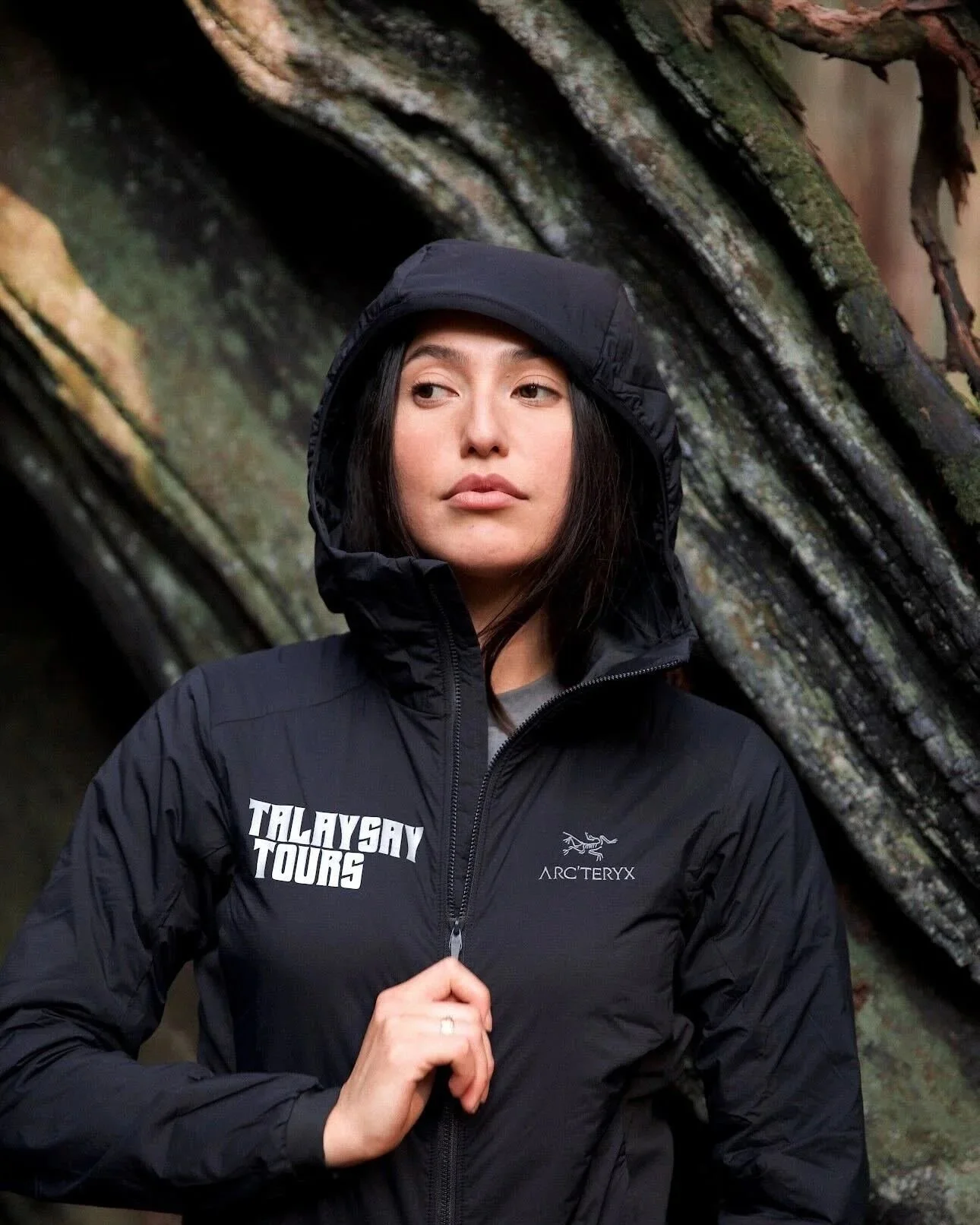 A woman wearing a black Arc'teryx jacket with a hood, standing outdoors near a rocky background, looking off to the side.