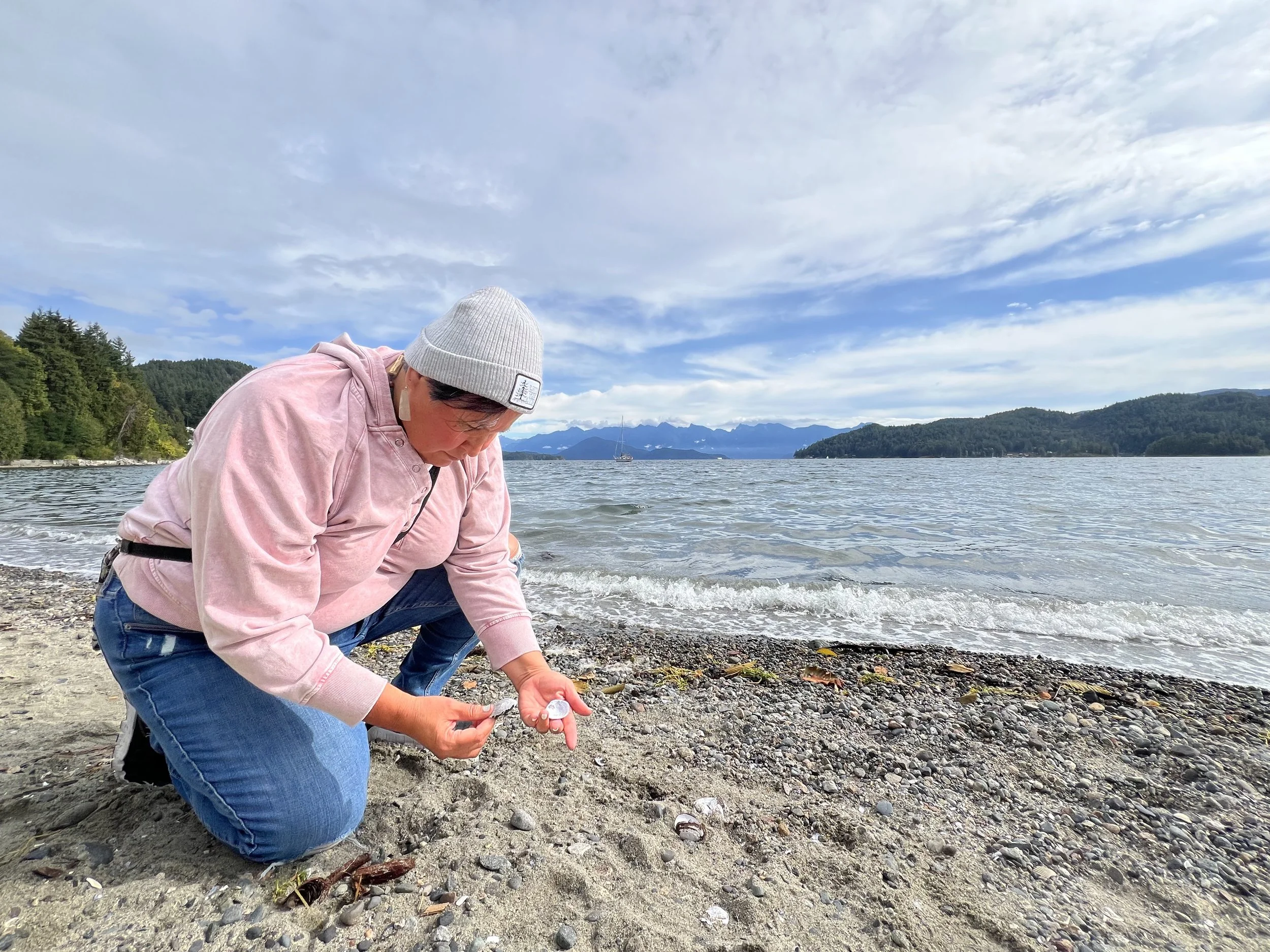 Person wearing pink hoodie and gray beanie collecting shells on a rocky beach by the water with mountains in the background.