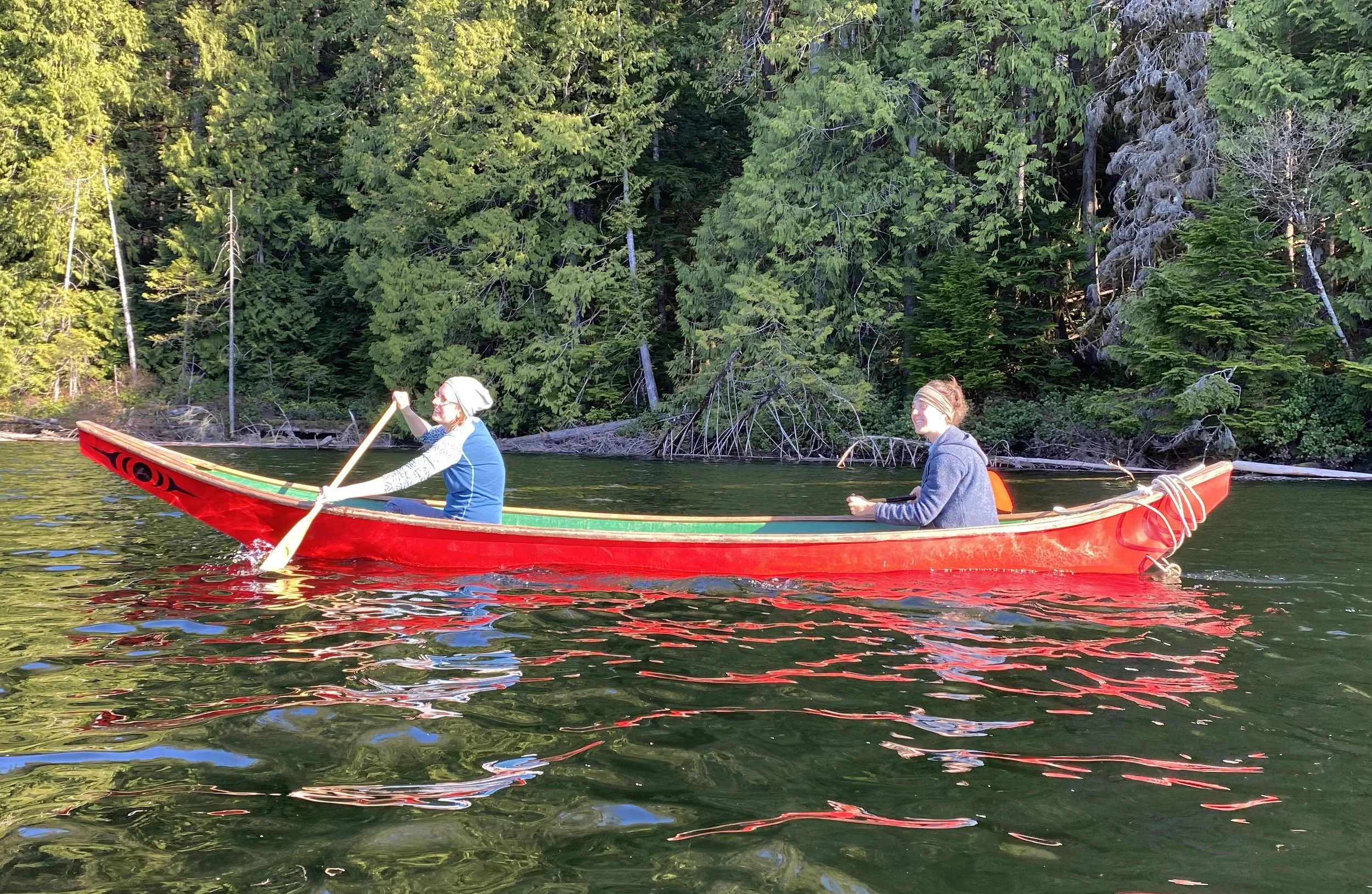 Two women paddling a red canoe on a lake surrounded by green trees.
