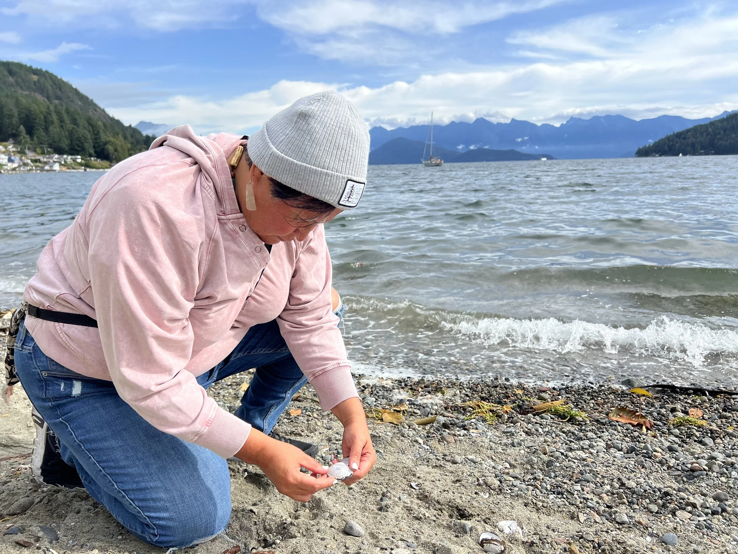 A person crouching on a pebble beach by a lake, picking up a seashell. The person is wearing a pink hoodie, blue jeans, and a light gray beanie. In the background, there are mountains, a sailboat on the lake, and a partly cloudy sky.