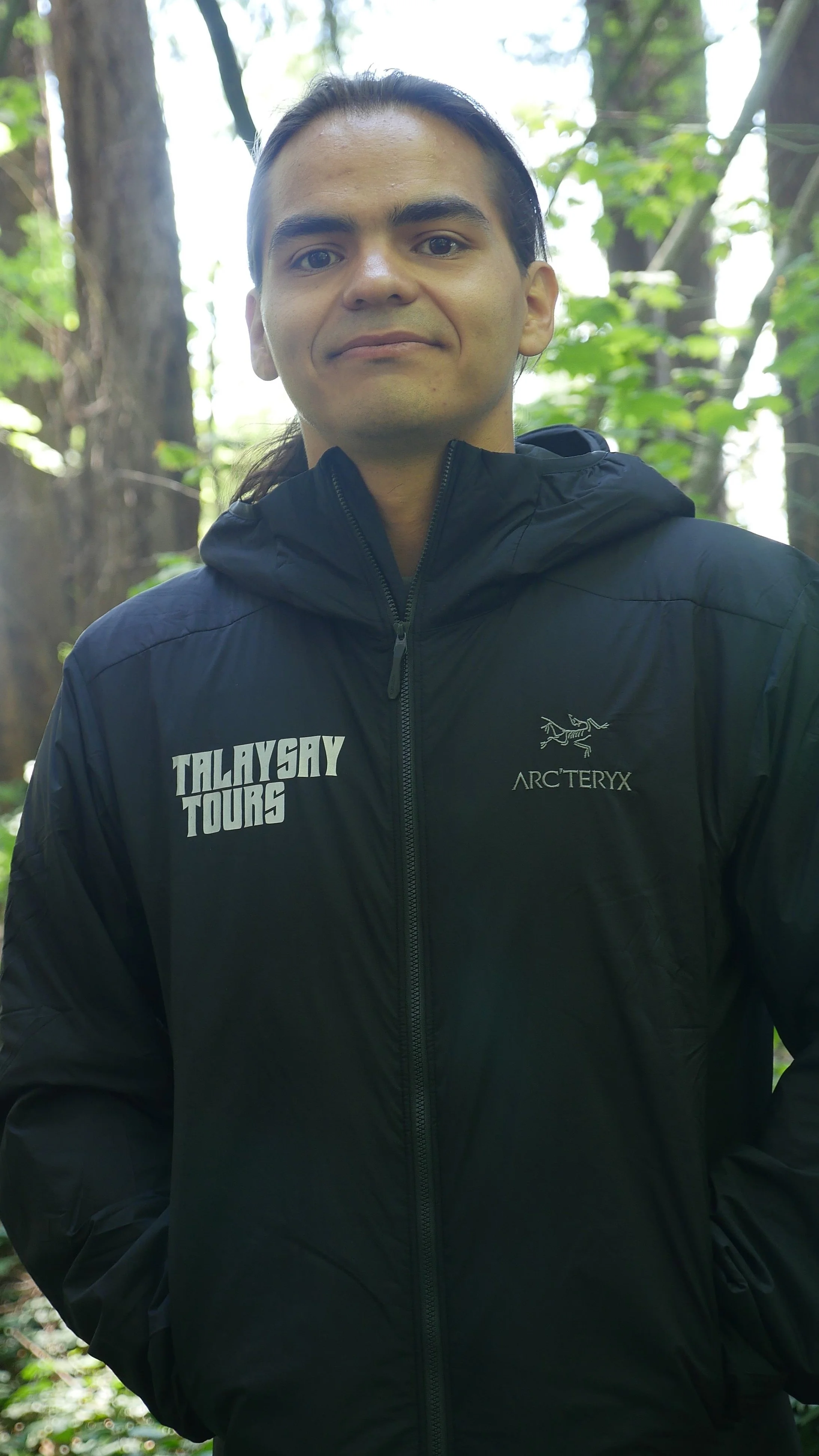 A man standing outdoors in a forest, wearing a black Arc'teryx jacket with 'TALAYSAY TOURS' printed on it.