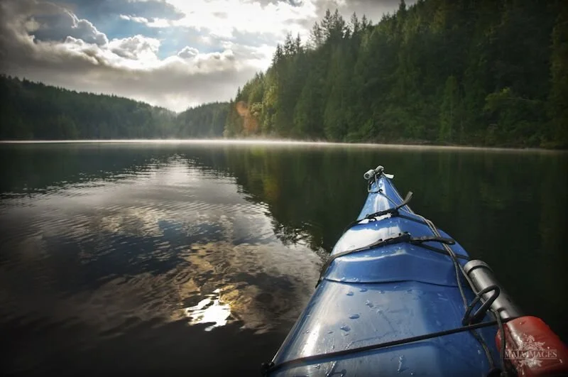 Photo from a kayak on a calm lake surrounded by forested hills under a cloudy sky.