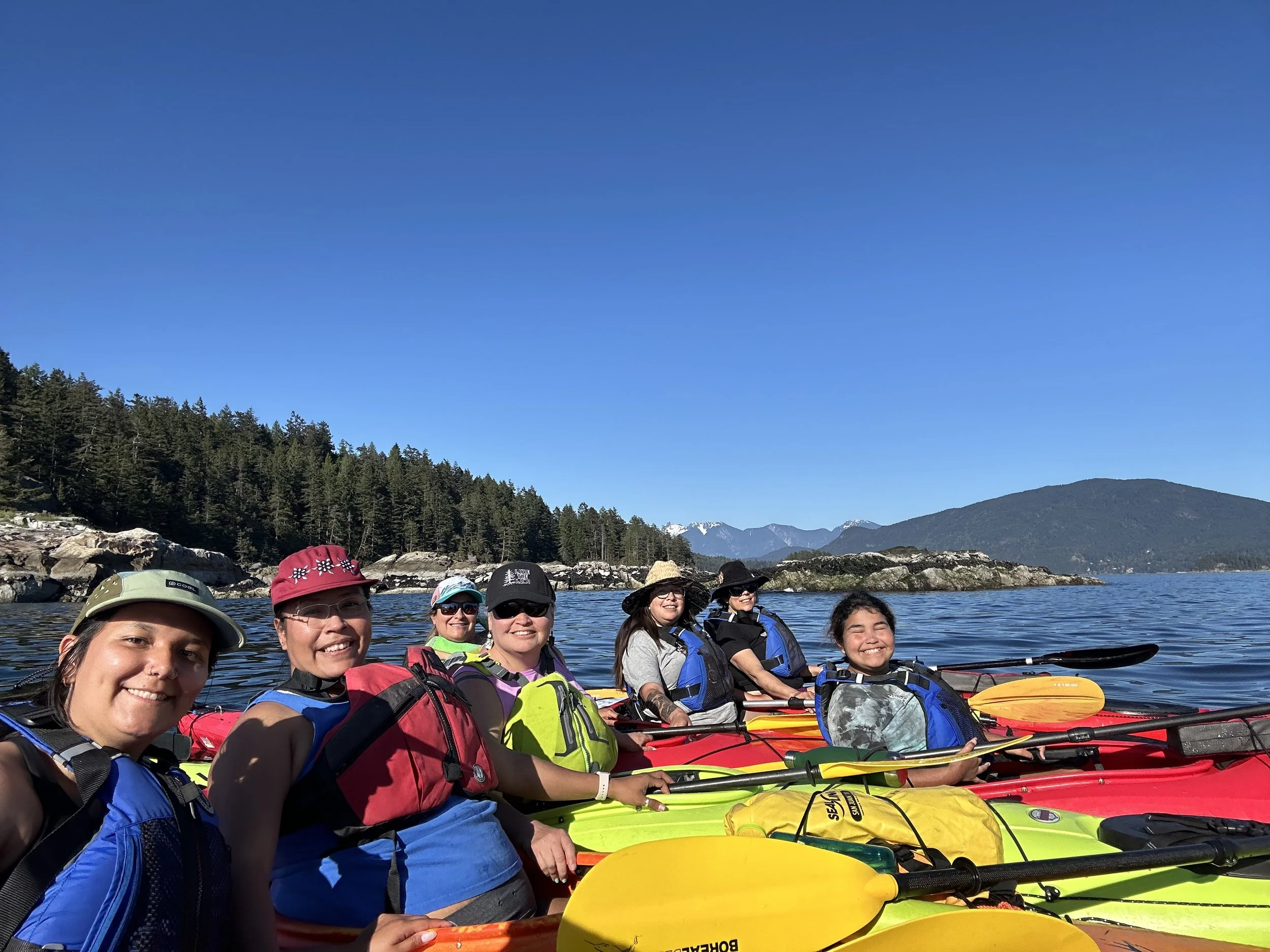 Seven women in kayaking gear smiling with kayaks on a lake, with trees and mountains in the background under a clear blue sky.