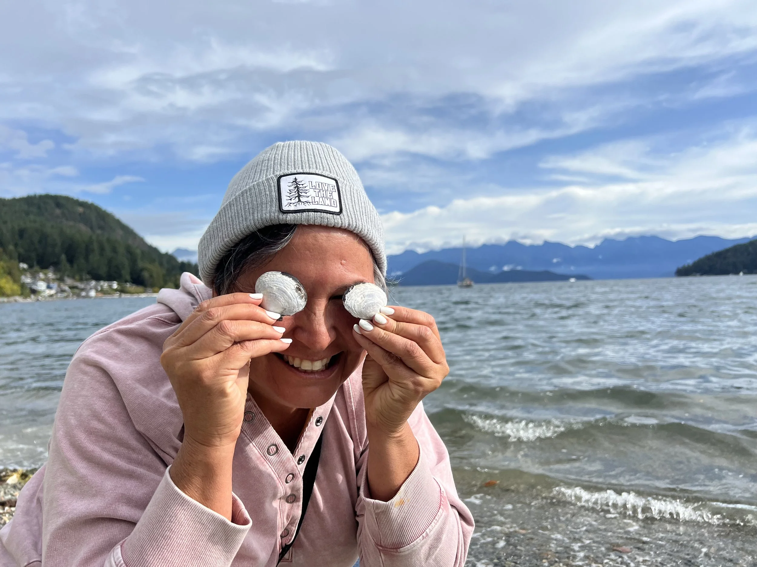 A smiling woman in a gray beanie and pink hoodie is playing with foiled seashells held over her eyes at a lakeside with mountains and a sailboat in the background.