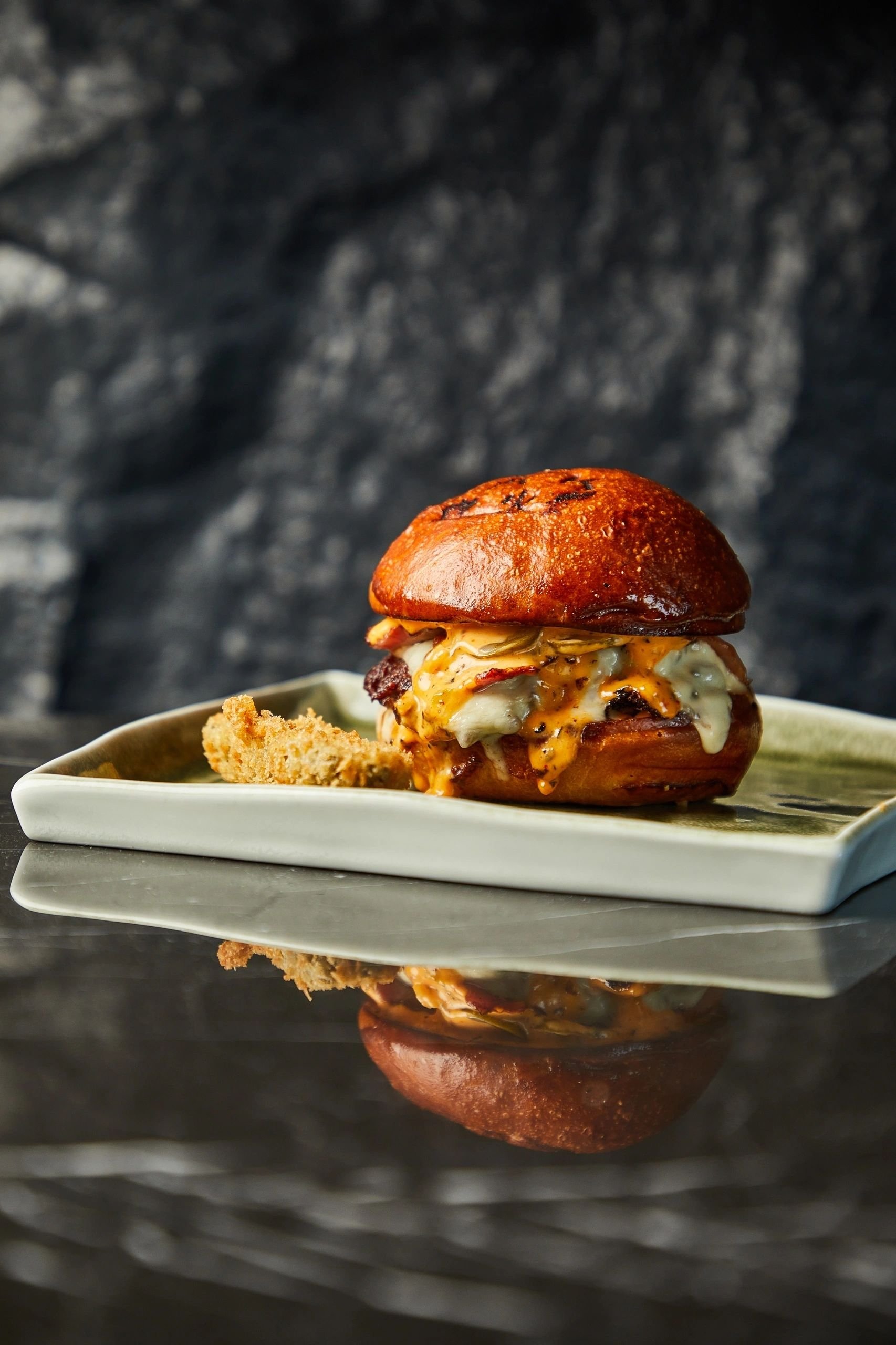 Gourmet cheeseburger with bacon and sauce on a brioche bun served on a square plate, dark background. F+B Strategy & Concepts, Branding & Collateral Design and Uniform Design by LEVELS creative studio in collaboration with Chef Marwan Sardouk.