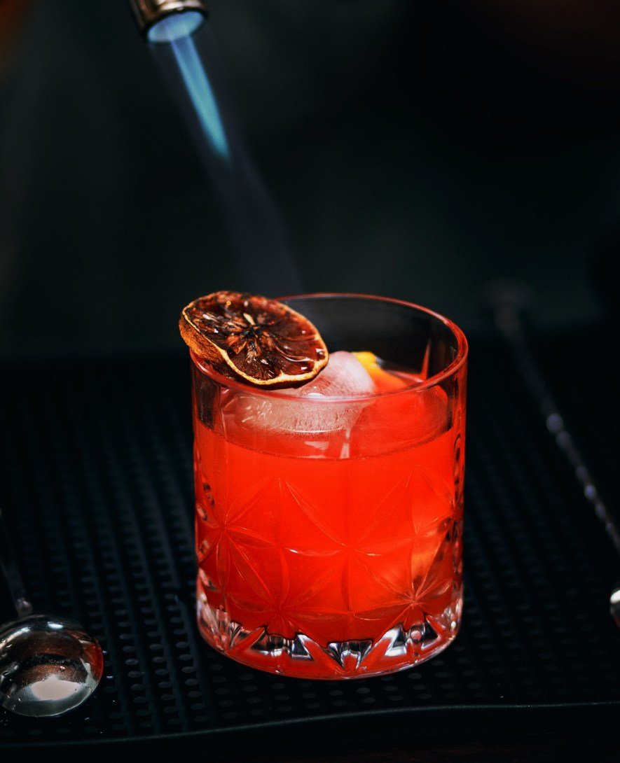 Red cocktail in glass with ice, garnished with dried citrus slice, torch flame above. Radisson RED Chandigarh, Mohali, India. F&B Concepts by LEVELS creative studio.