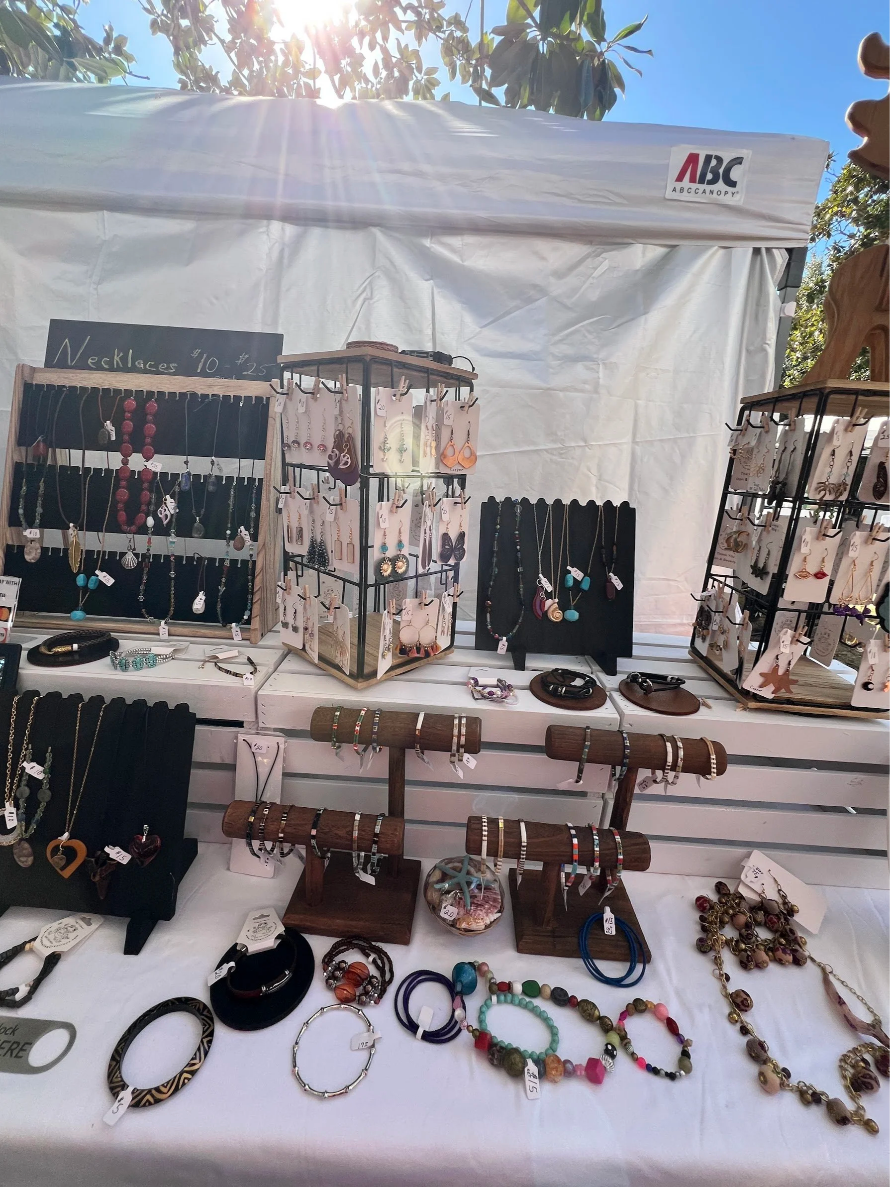Handcrafted artisan jewelry booth display featuring handmade necklaces, beaded bracelets, gemstone earrings, and unique statement pieces showcased on wooden stands at an outdoor market.