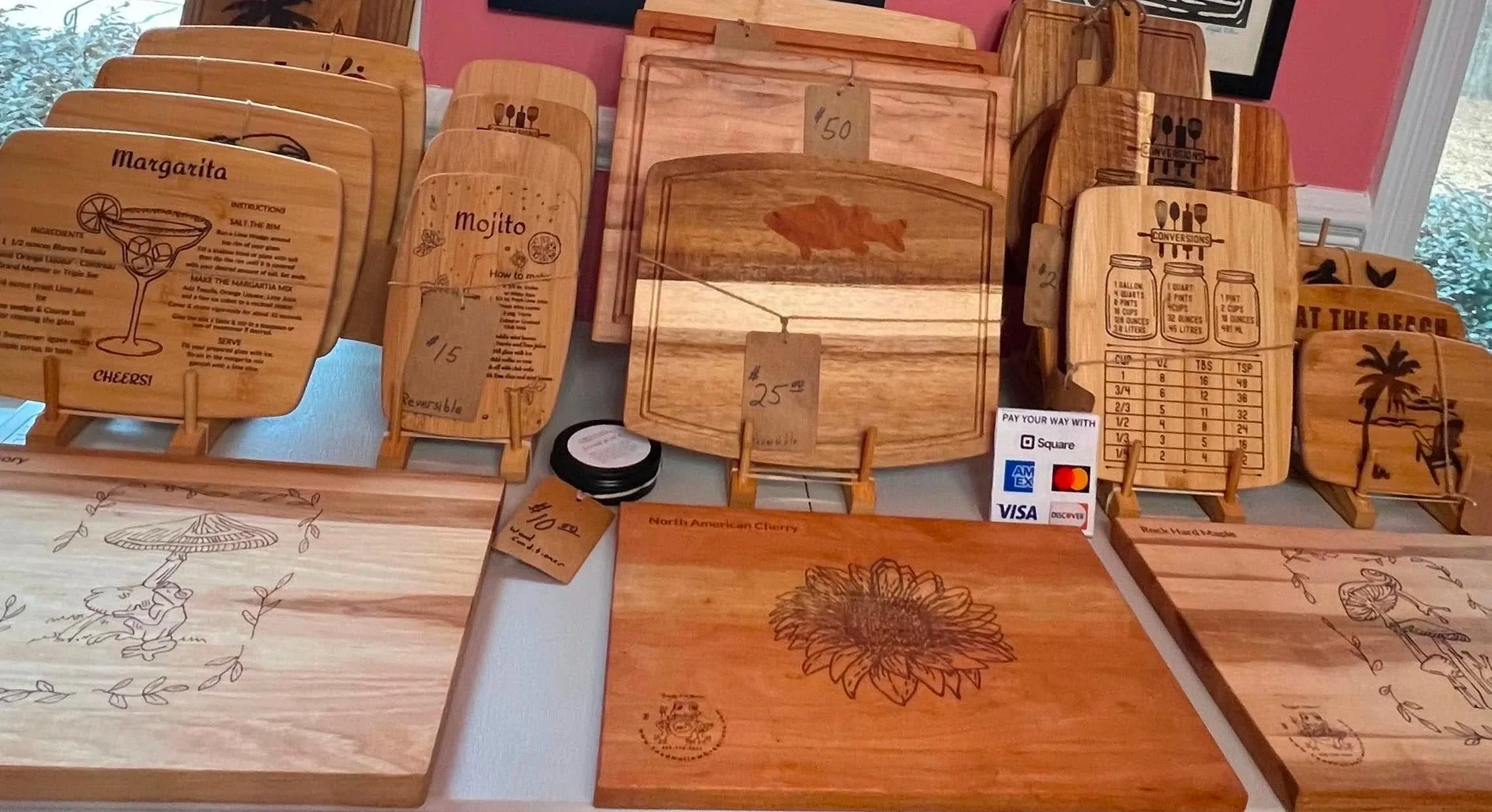 Handcrafted wooden cutting boards and engraved serving boards featuring beach-themed and cocktail designs displayed at an outdoor artisan market booth, including charcuterie boards and custom wood kitchen décor.