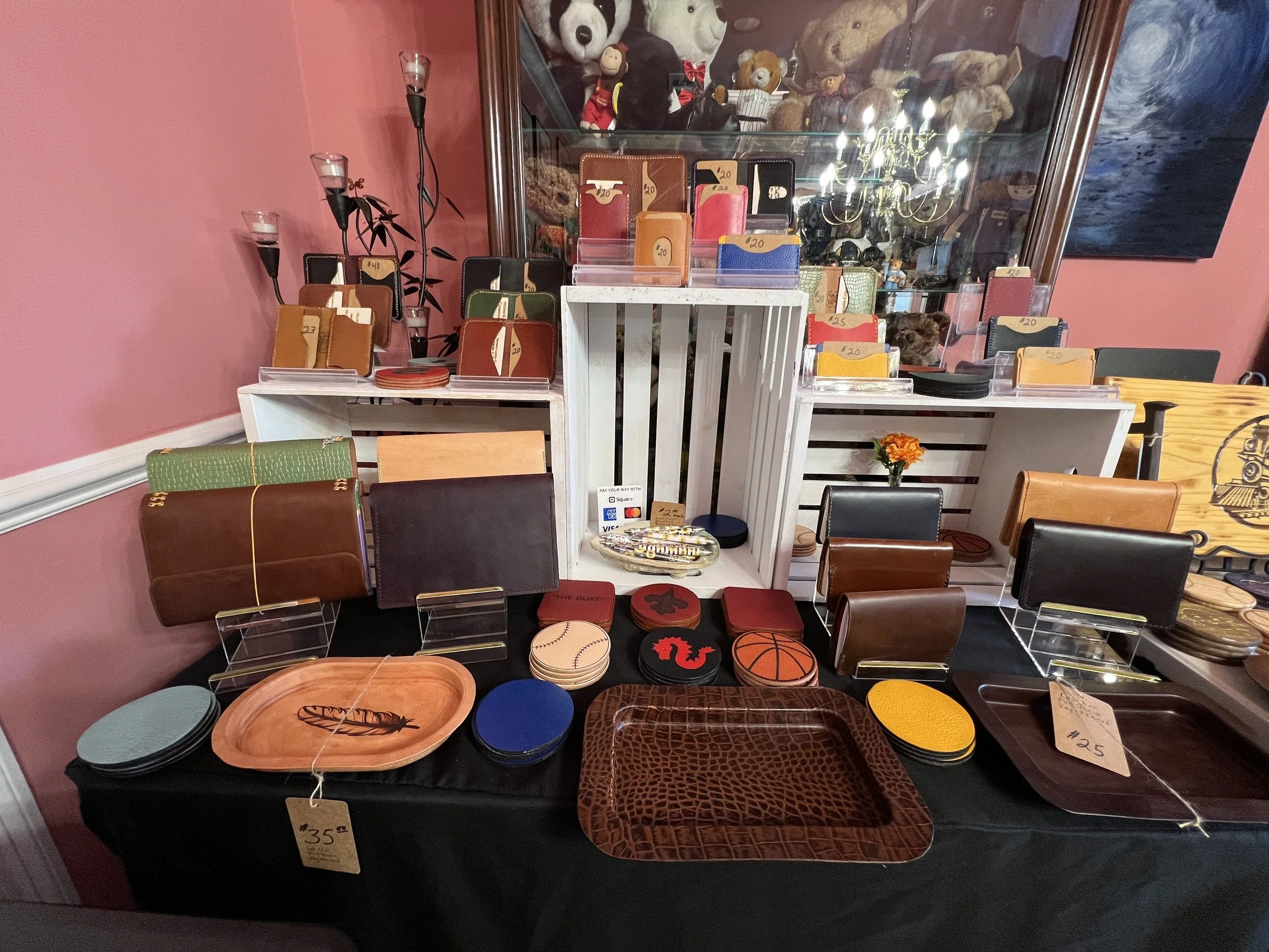 Handcrafted full-grain veg-tanned leather wallets, minimalist card holders, journals, coasters, and EDC valet trays displayed at artisan booth featuring premium leather goods in brown, tan, red, and mustard finishes.
