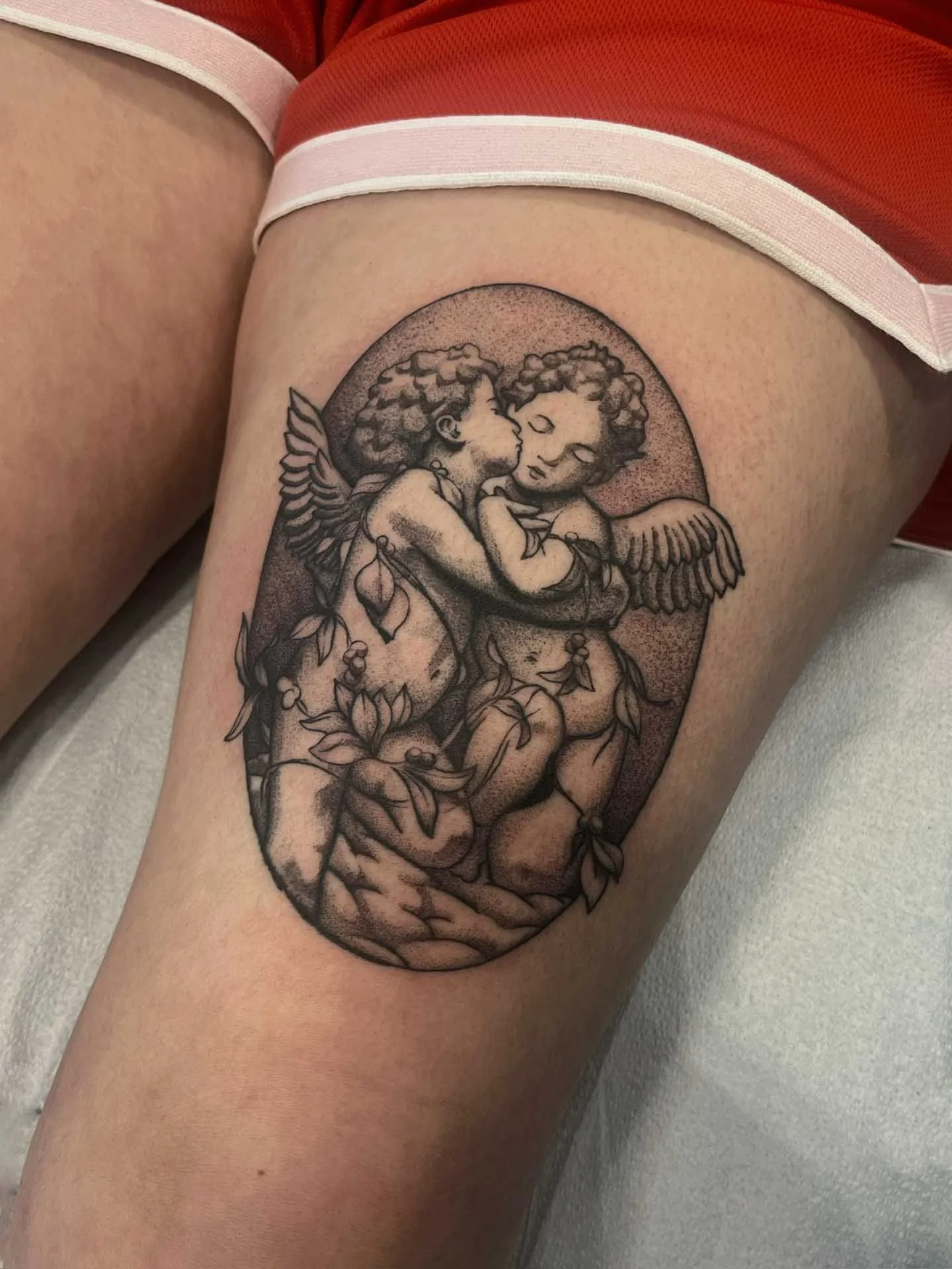 Surreal black and gray tattoo by @kuya.tattoos of two cherubs embracing and kissing, with wings and surrounded by flowers, within an oval frame. Kuya tattoos at Dark Water, East Bay's top rated tattoo studio.
