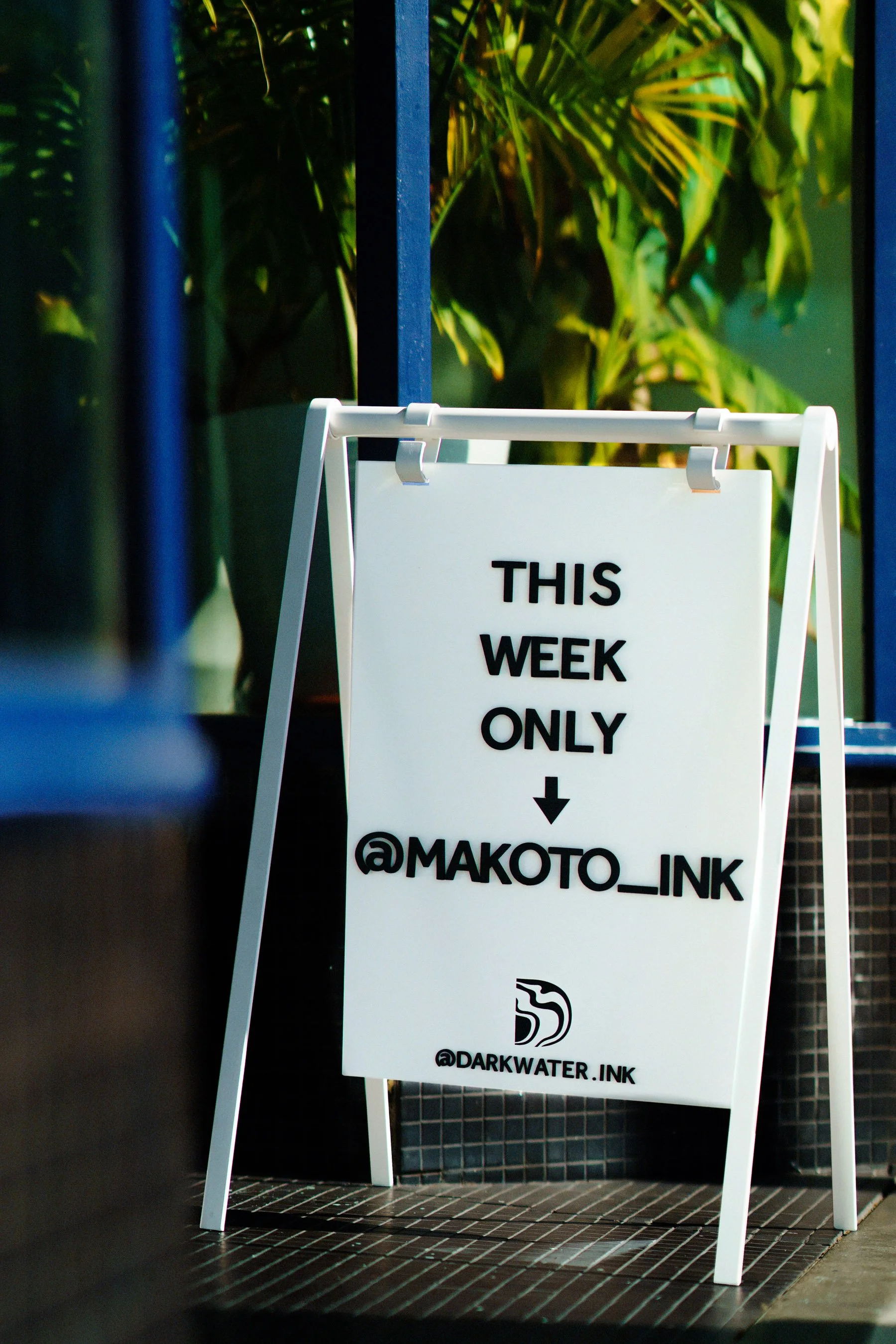 White A-frame signboard with black text that says 'This week only' and Instagram handles '@makoto_link' and '@darkwater.ink', set outside with green plants in the background.