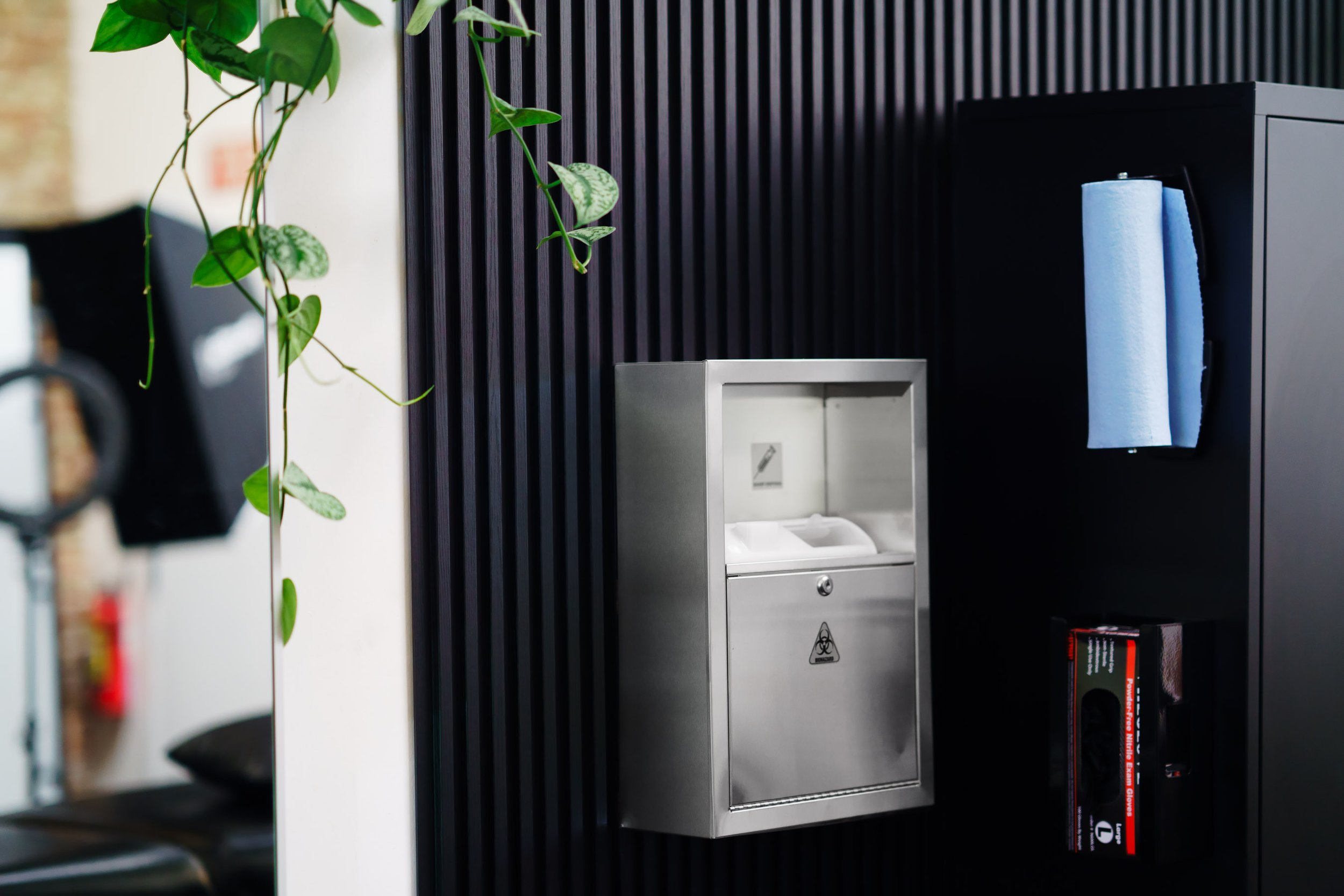A stainless steel biohazard disposal bin mounted on a black wall, with a paper towel dispenser nearby and green plant leaves hanging in the foreground.