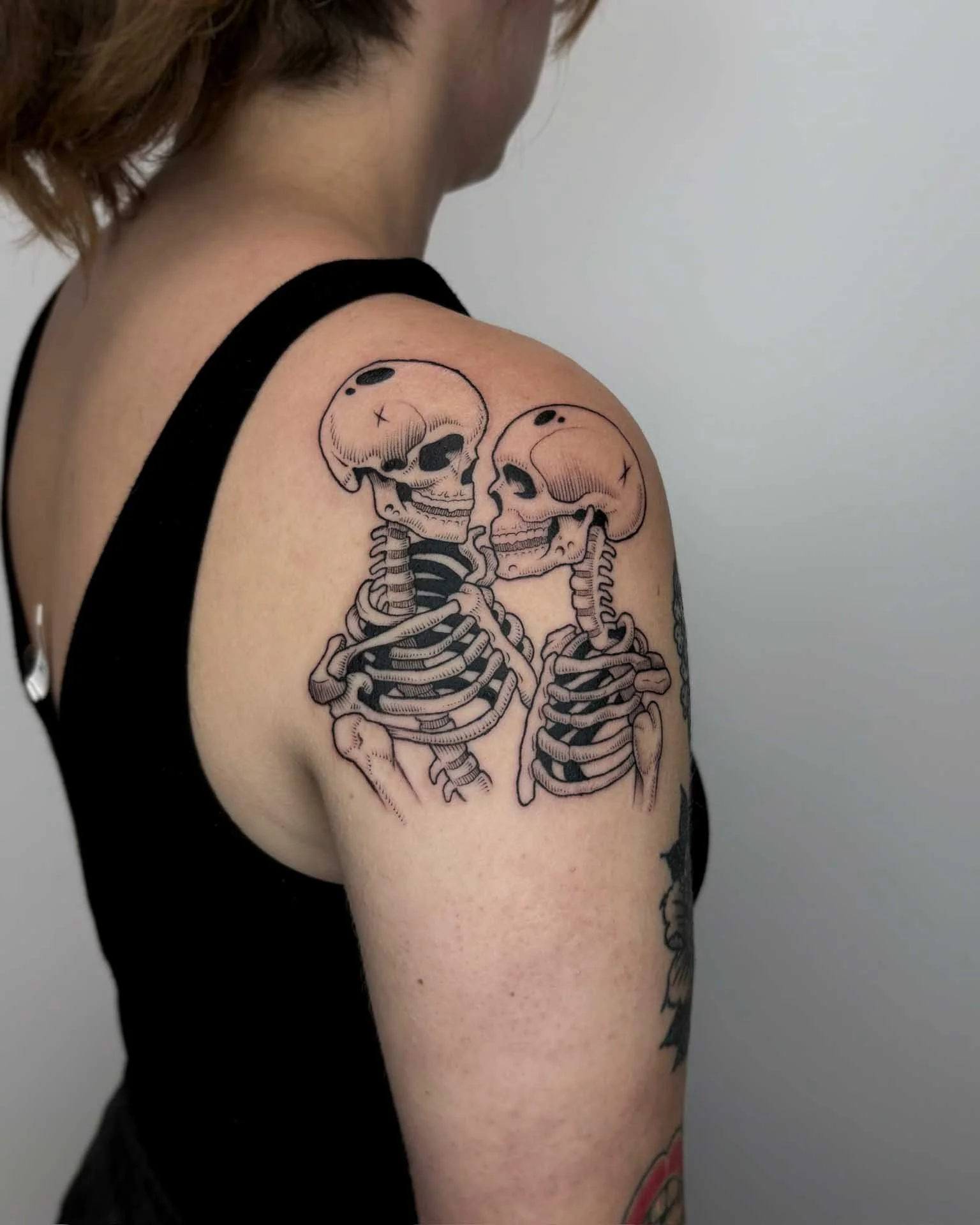 Surreal tattoo by Kuya (@kuya.tattoos)of two skeletons with skulls touching on a person's upper arm and shoulder at top-rated Berkeley studio, Dark Water.