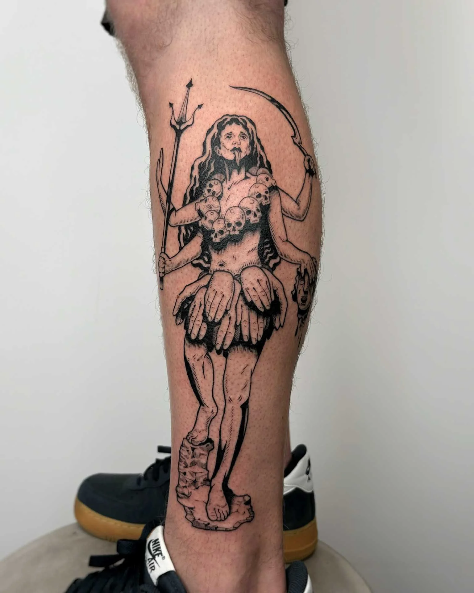 Surreal tattoo by Kuya (@kuyatattoos) of a deity with multiple arms, holding a trident and a curved blade, with a crown of skulls around her neck, standing on a pedestal. Find Kuya at Dark Water, Berkeley's top-rated tattoo studio.