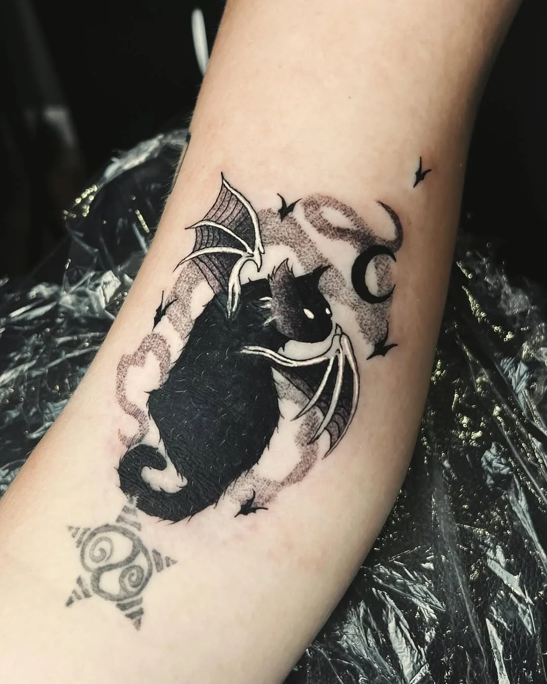 Black cat with bat wings. Witchy black and grey tattoo of a vampire cat under a crescent moon, with dotwork shading and dancing bats. By Stella Sanchez (@umbra.inks), now booking at Dark Water Tattoo Studio in Berkeley, CA.