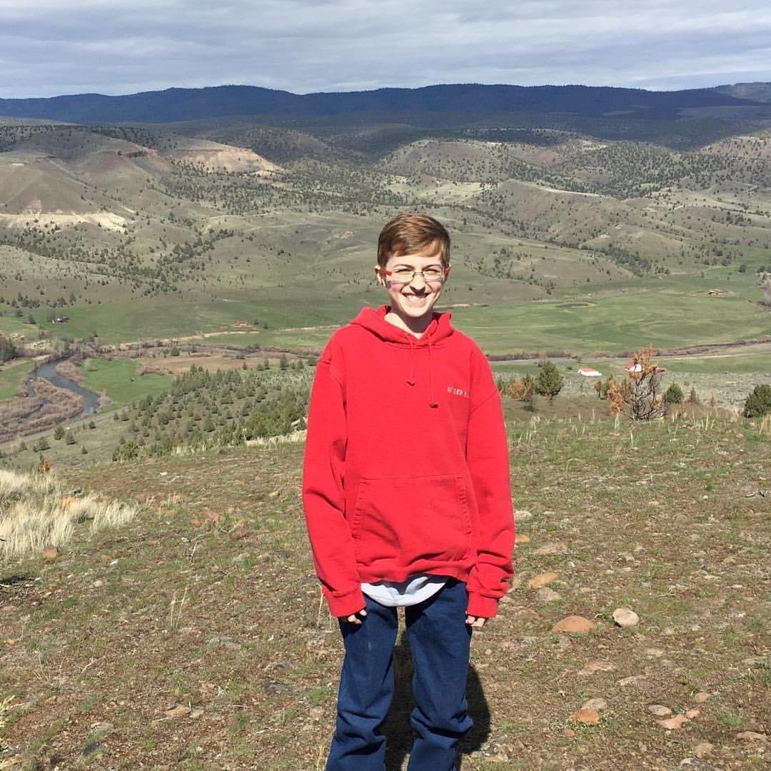 A boy in a red hoodie and glasses smiling outdoors on a hilltop with rolling mountains and green fields in the background.